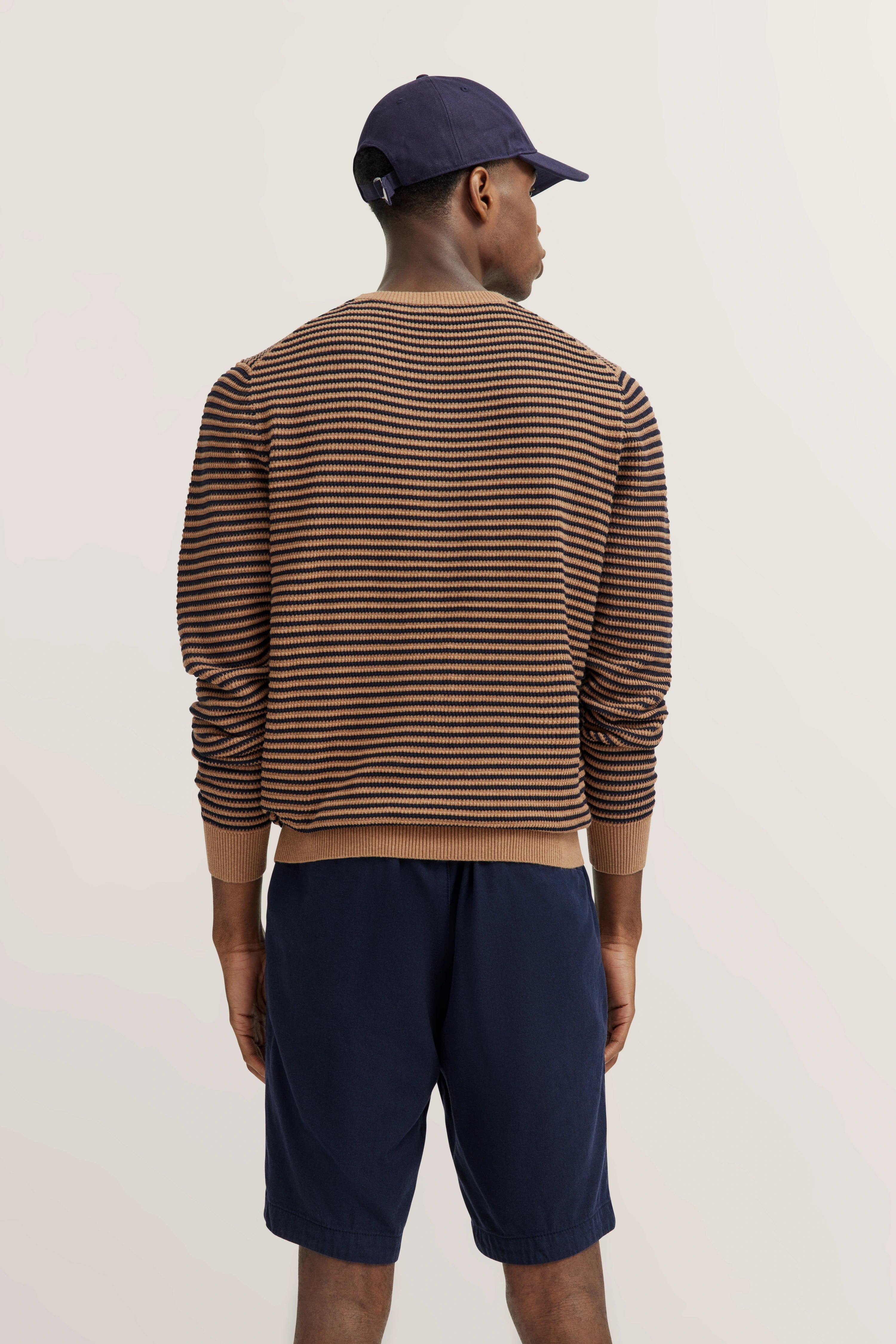 Modell in gestreiftem Pullover, braun-blau, mit dunkelblauer Shorts und Cap, Rückansicht, entspanntes Posing.