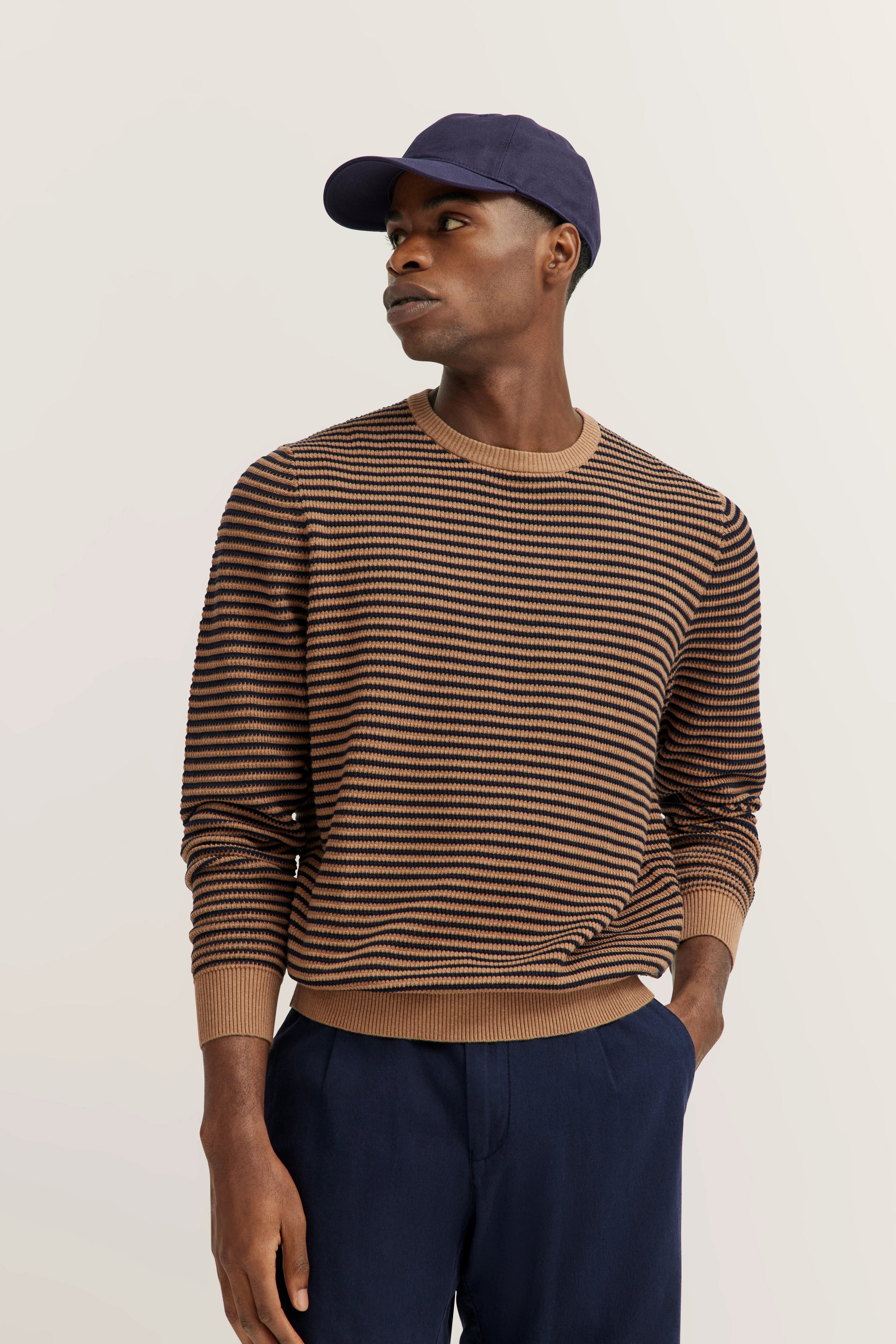 Model in gestreepte trui, bruin en donkerblauw, met blauwe pet. Casual, zijwaartse blik.