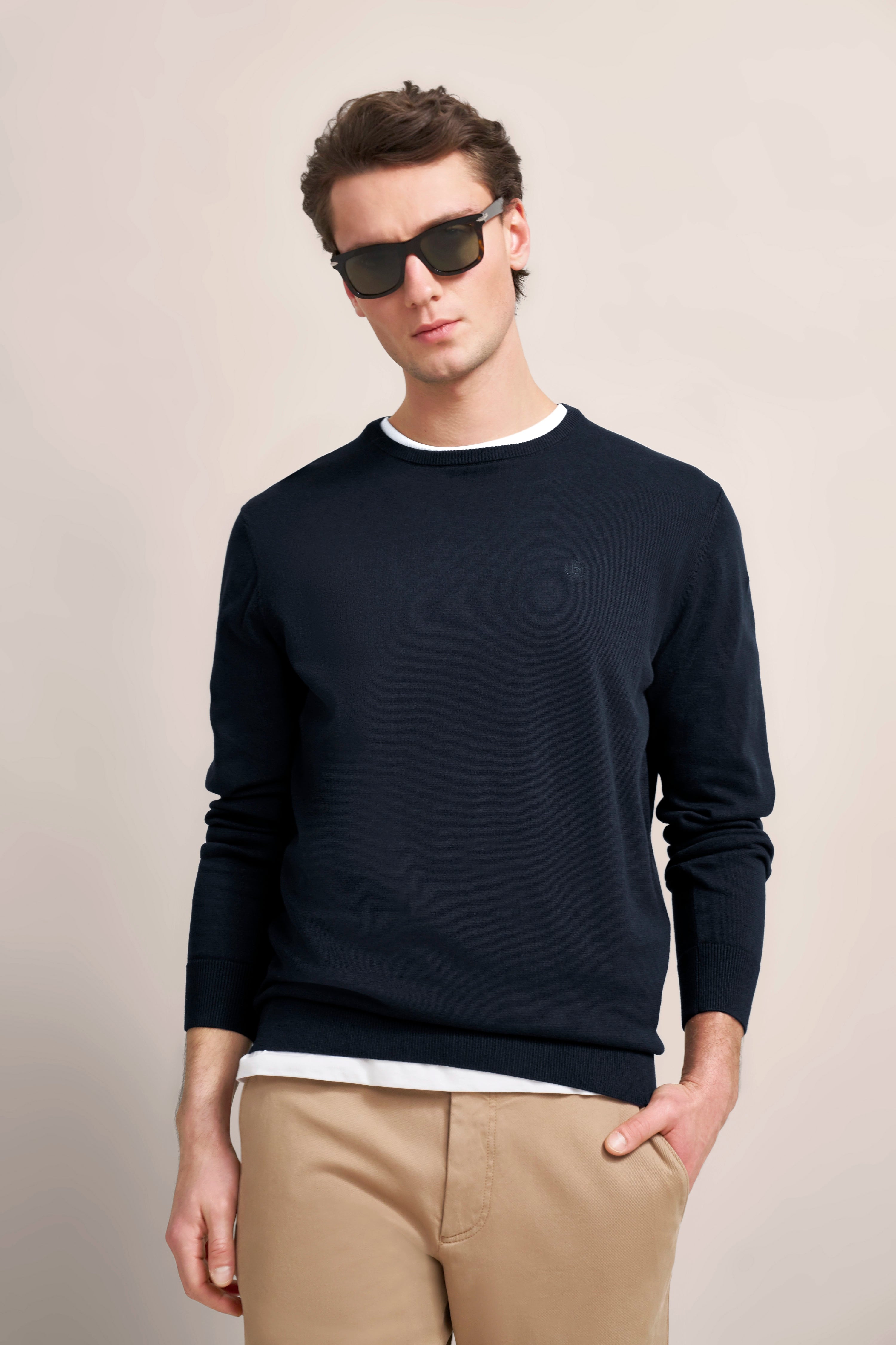bugatti Modelbild: Dunkler Pullover, beige Hose, weißes Shirt. Lässig mit Sonnenbrille, entspanntes Posing.