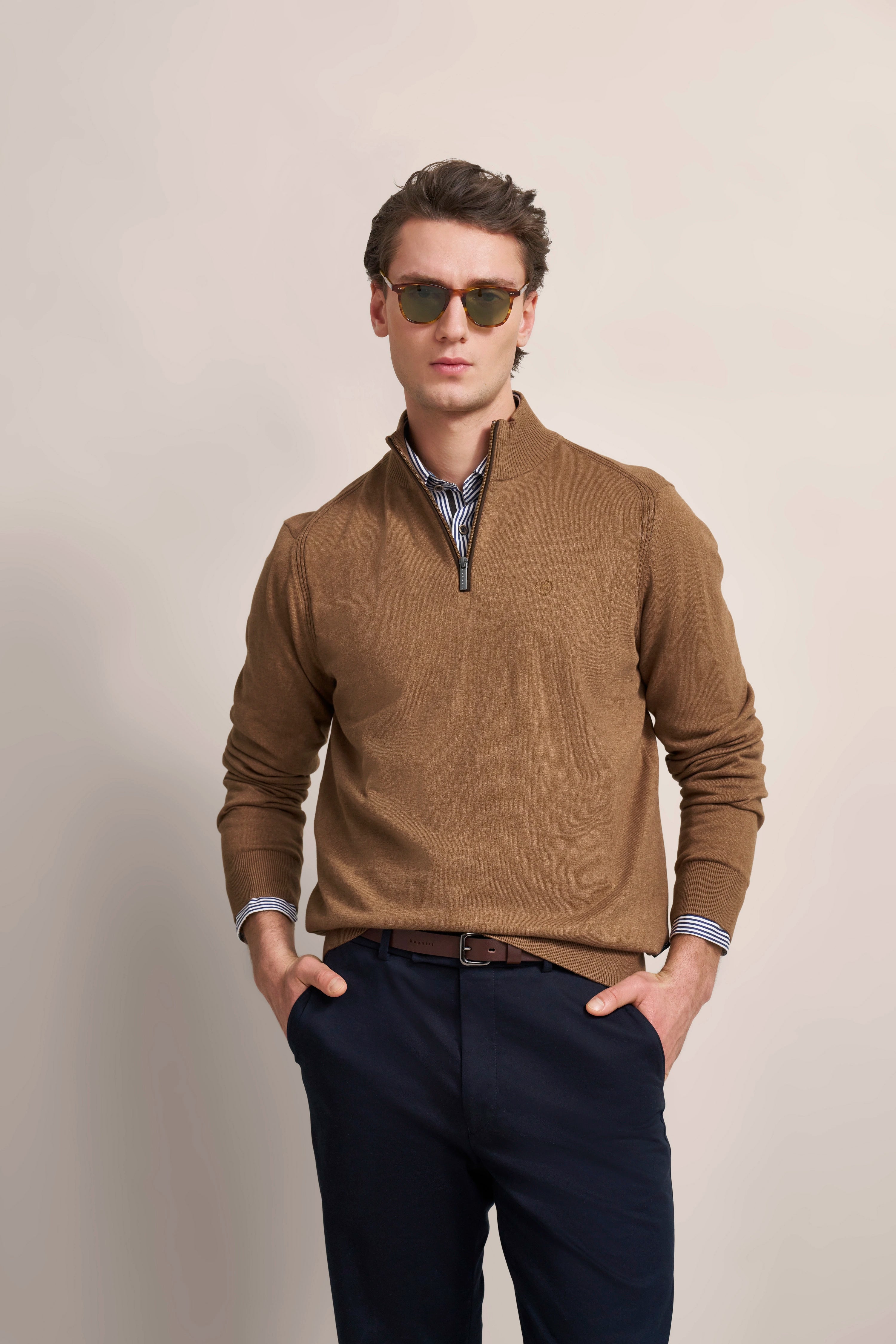 Model in braunem Pullover und blauer Hose, Hände in Taschen, mit Sonnenbrille.