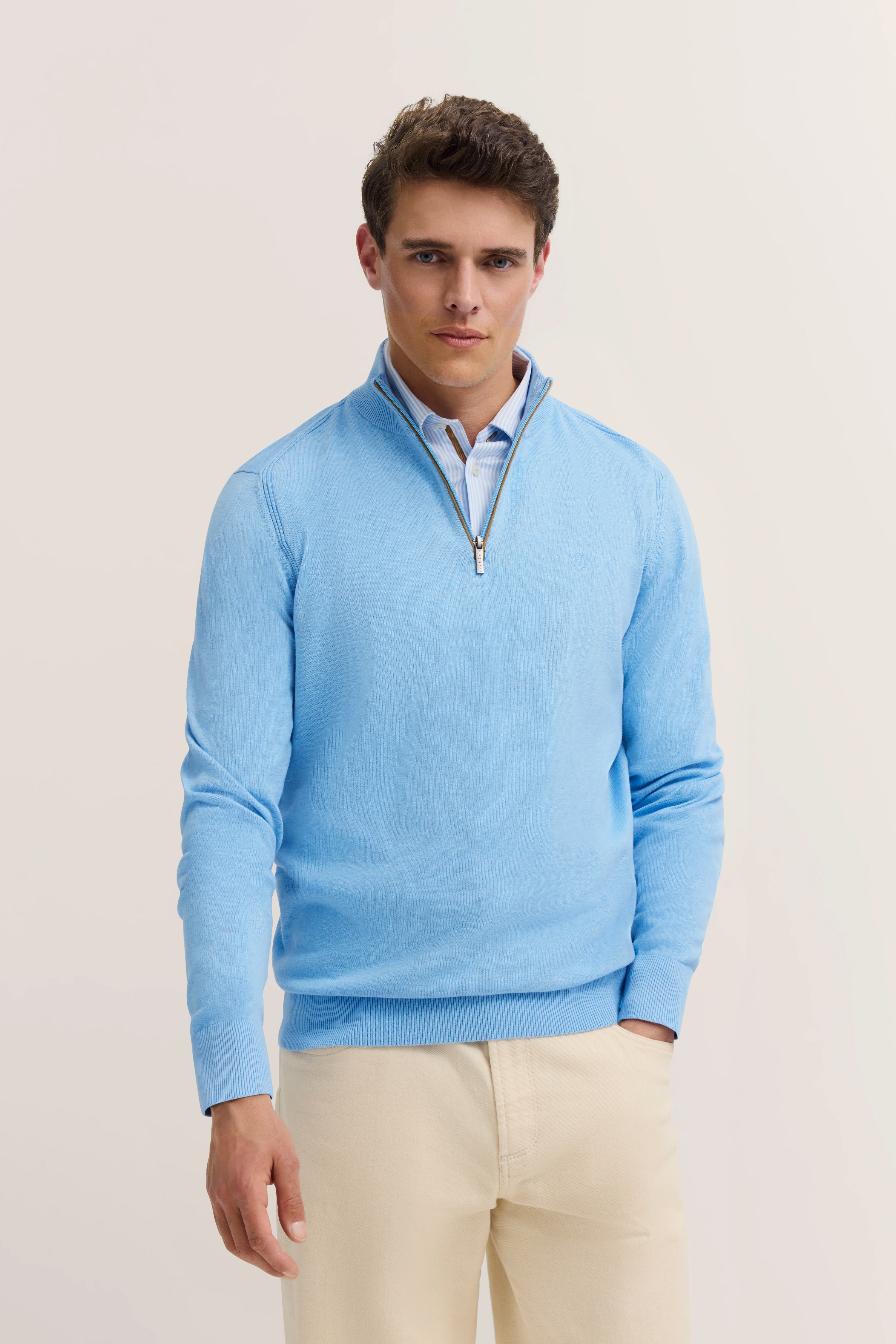 Model in lichtblau bugatti trui, beige broek, ontspannen pose, stijlvolle uitstraling.