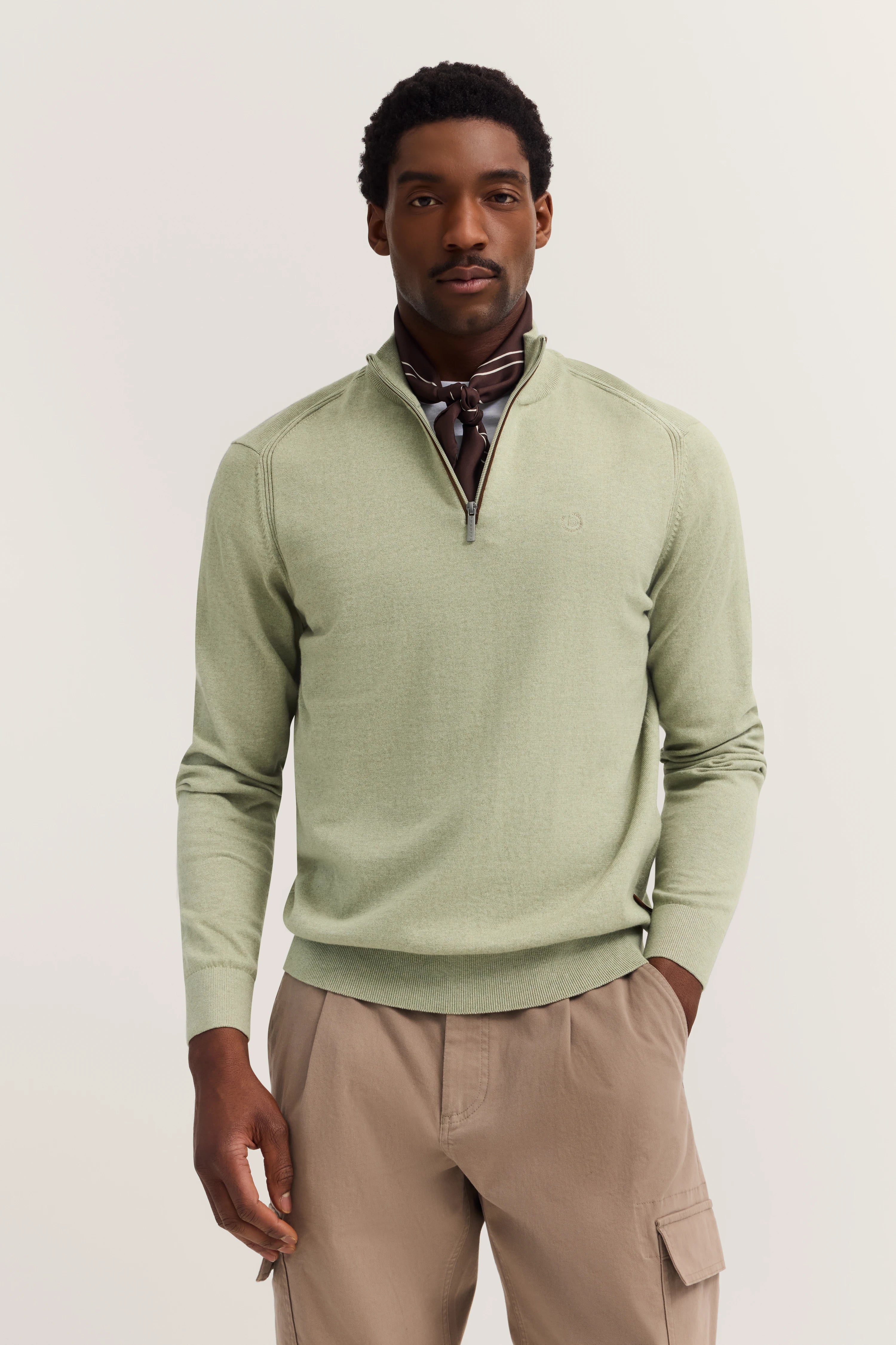 bugatti Modelbild: Mann in grünem Pullover, braunes Halstuch, beige Hose, entspannte Pose.