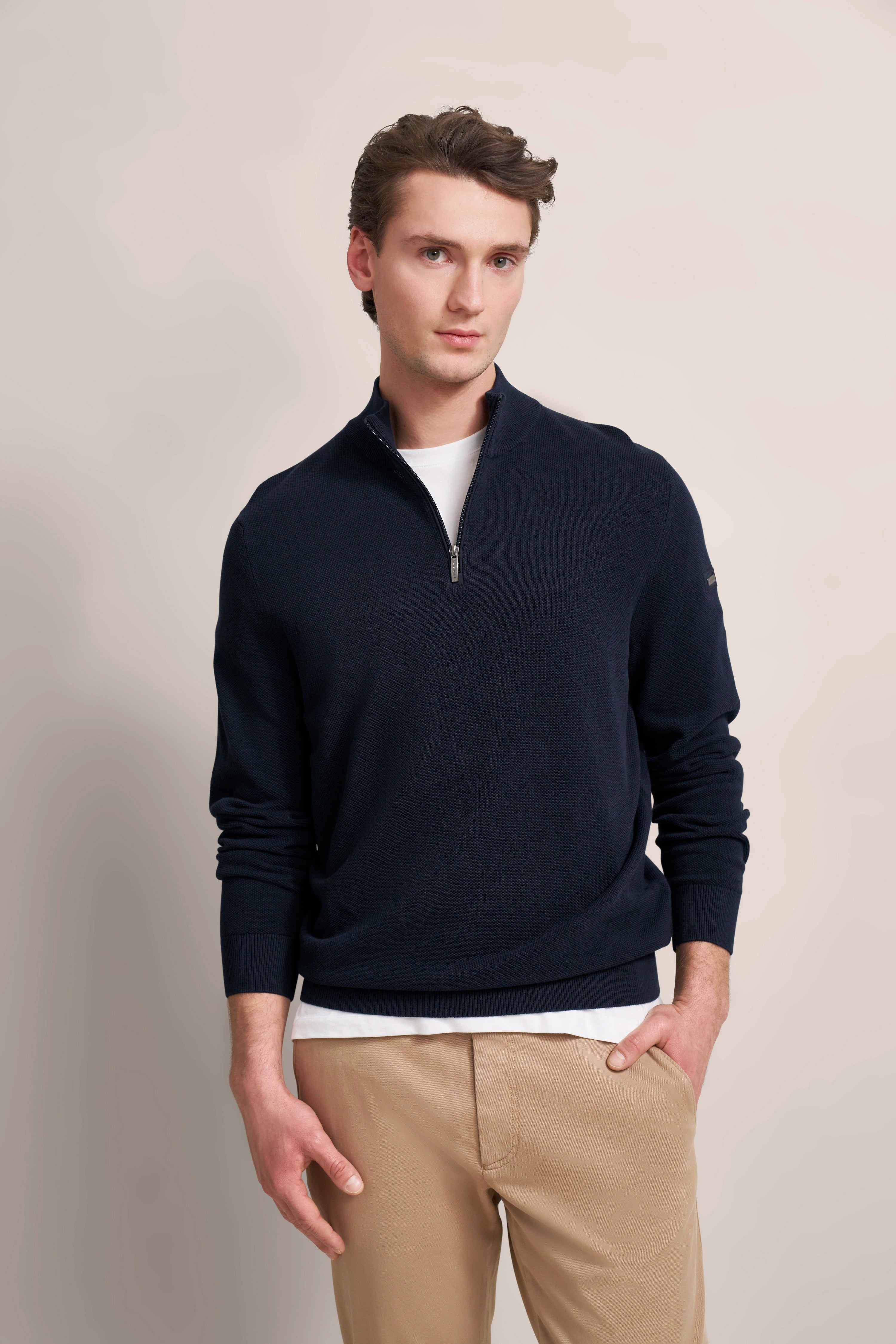 Model mit dunkelblauem bugatti Pullover, weißem Shirt und beigen Chinos. Lässige Pose mit entspanntem Ausdruck.