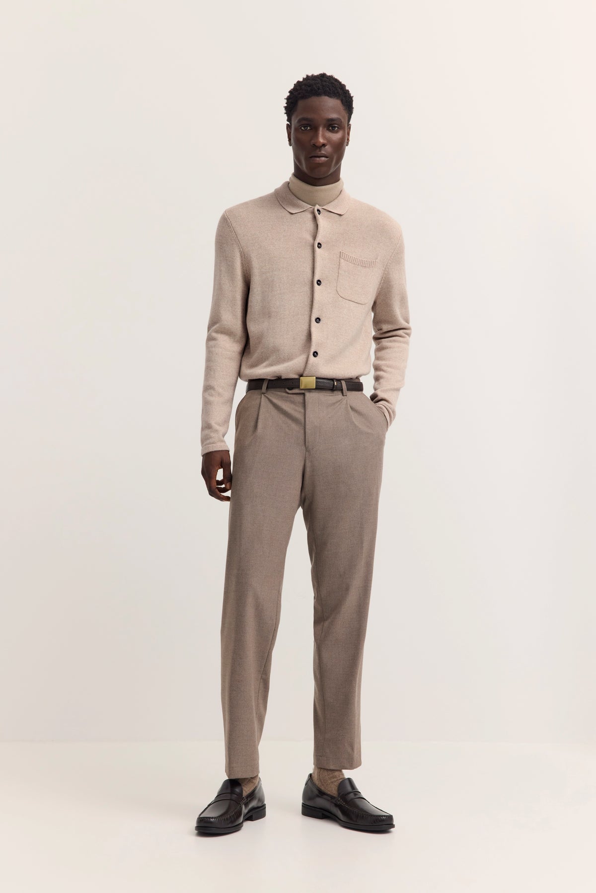 Beiges bugatti Outfit mit Strickpullover und Anzughose, Model steht lässig mit Händen in Hosentaschen.