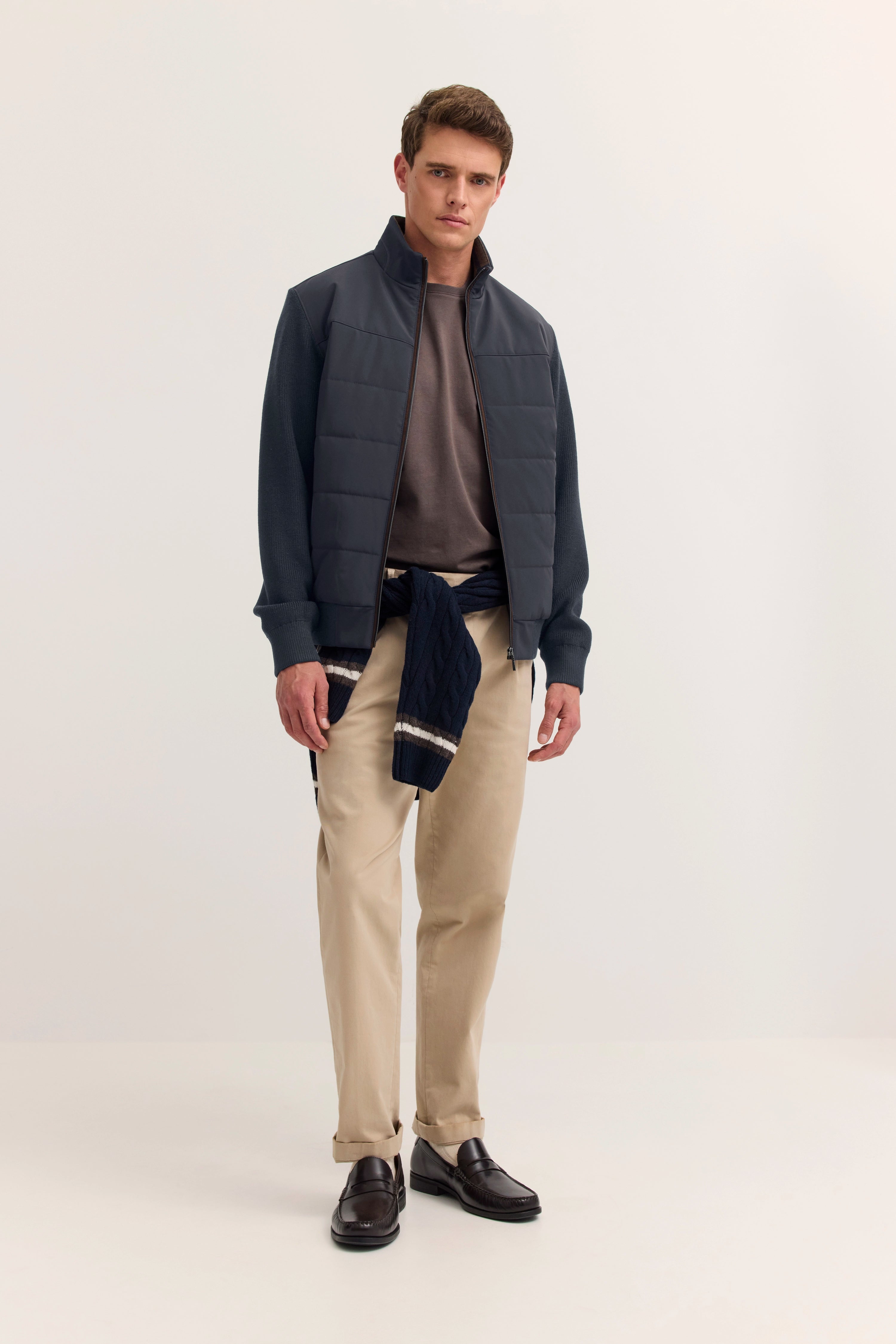 Model in navy Jacke, braunem Shirt, beigen Hosen und Loafern. Lässiges Stehen, Hände locker.