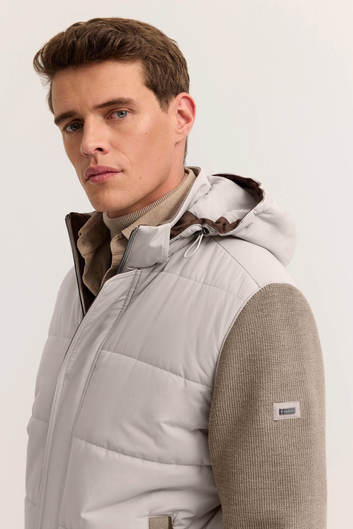 Model trägt bugatti Steppweste in Beige mit Kapuze, darunter beiger Strickpullover