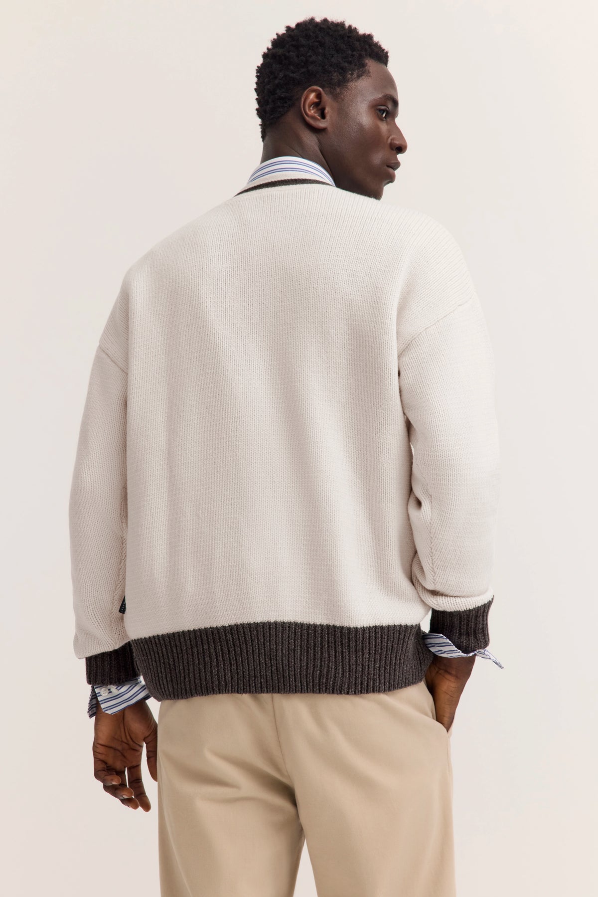 Model in cremefarbenem bugatti Strickpullover mit dunkelbraunem Saum, beiger Hose, Rückansicht.