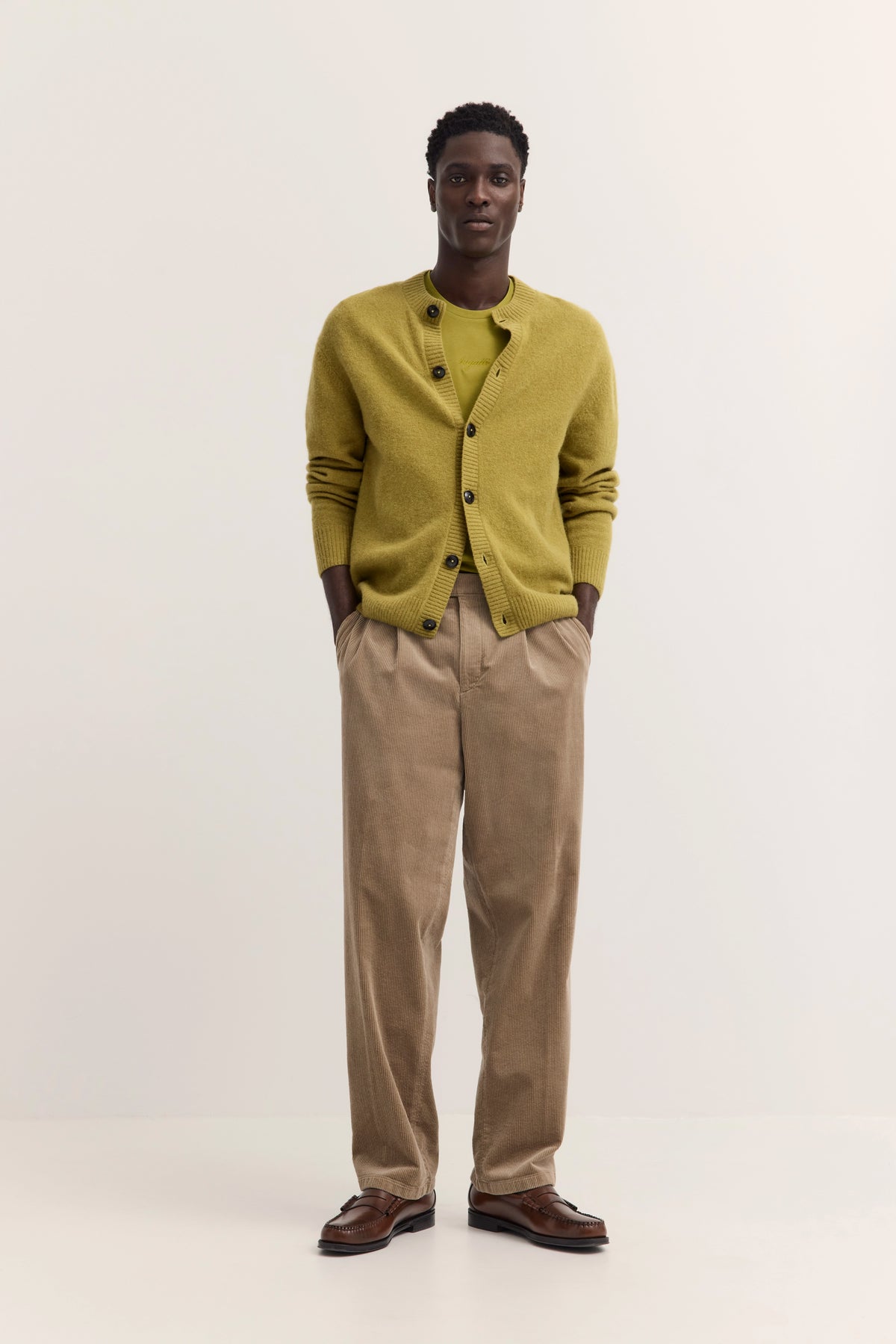 Model in olivgrünem Cardigan und Shirt, beige Hose, braune Schuhe, legeres Posing.