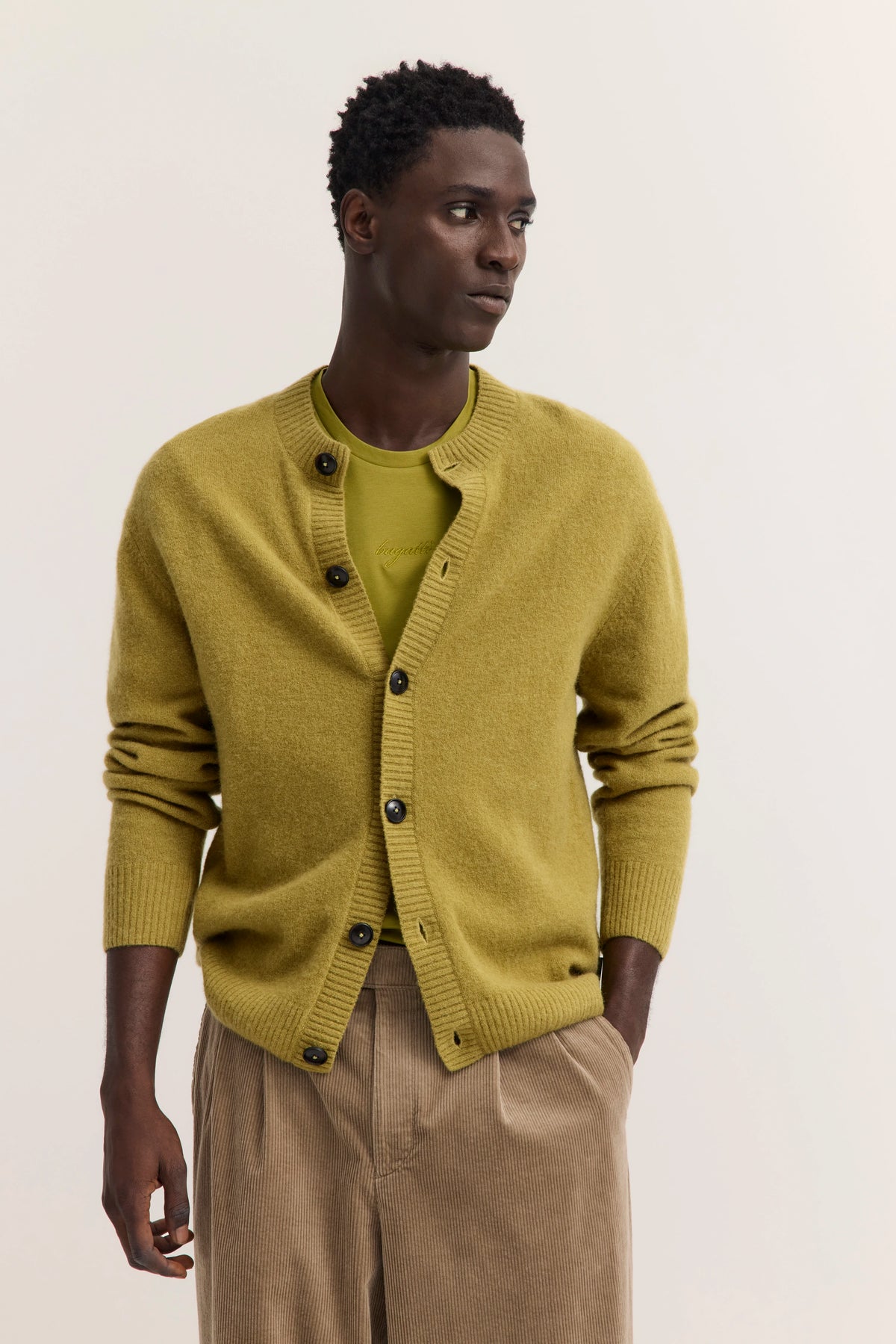 Model in olivgrüner bugatti Strickjacke und Shirt, beige Hose, entspanntes Posing.
