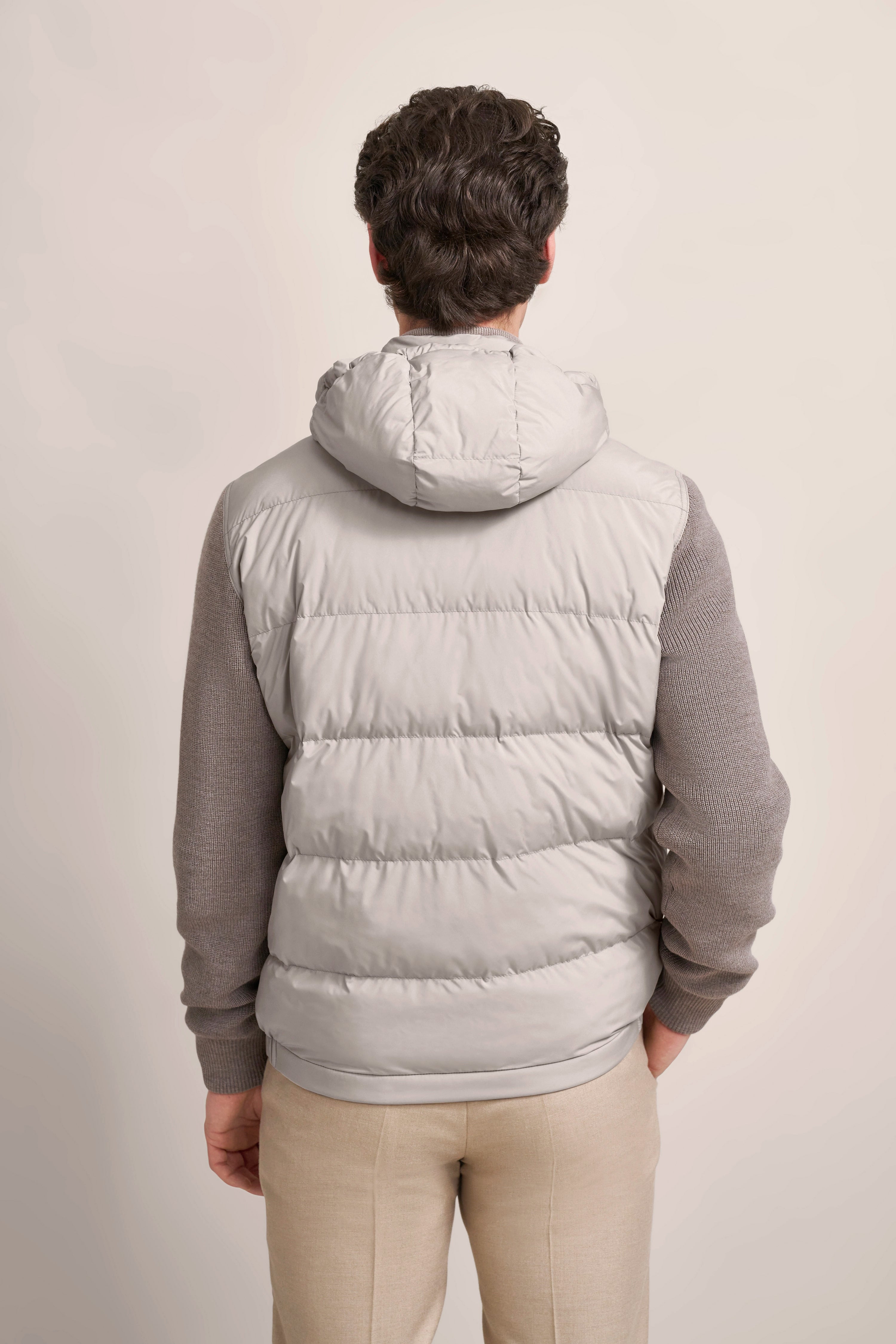 Rückansicht eines Models mit grauer bugatti Daunenweste, grauem Pullover und beiger Hose, lässig stehend.