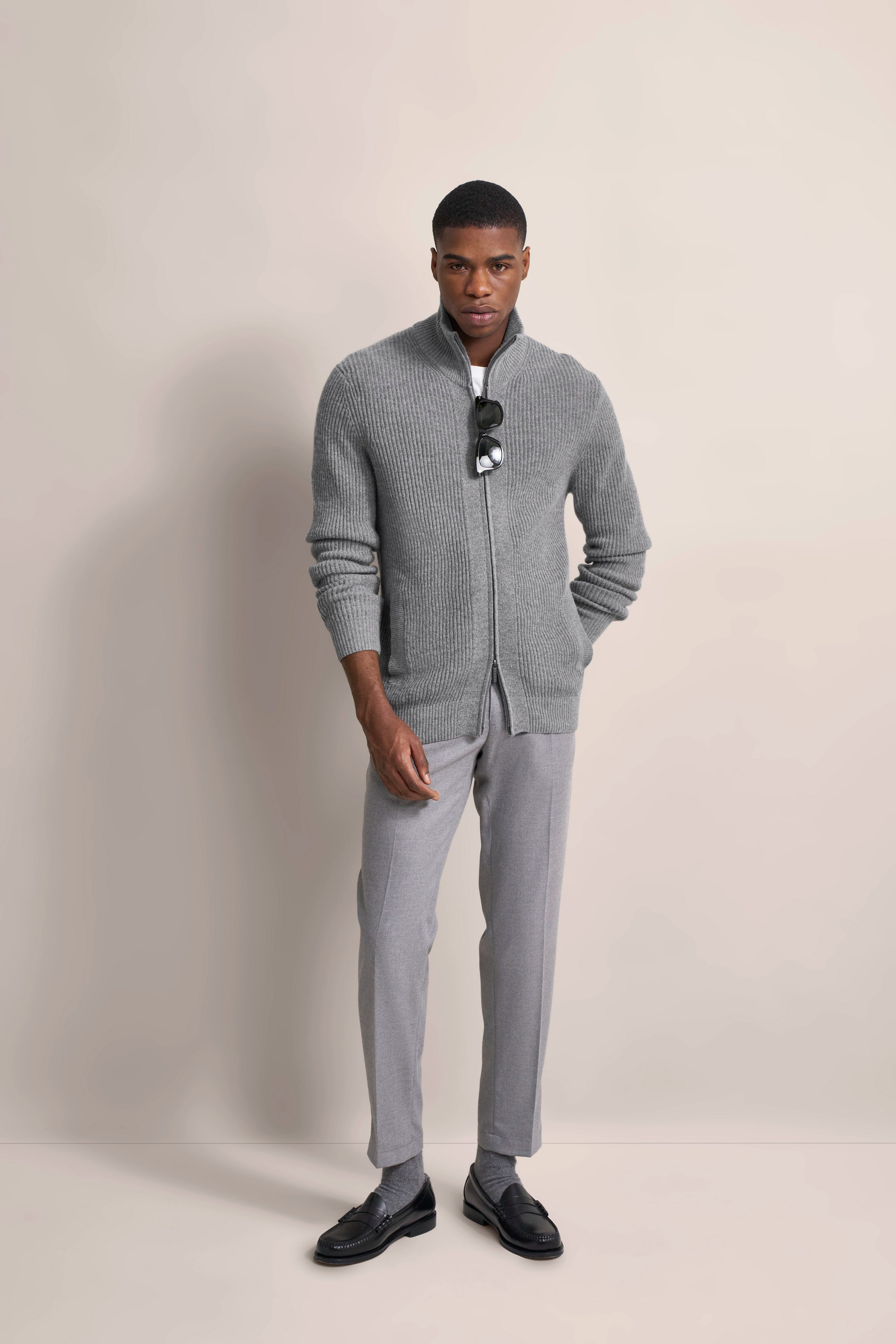 Grauer Strickpullover und Hose, Model posiert stehend mit Sonnenbrille in Hand, bugatti Stil und Eleganz.