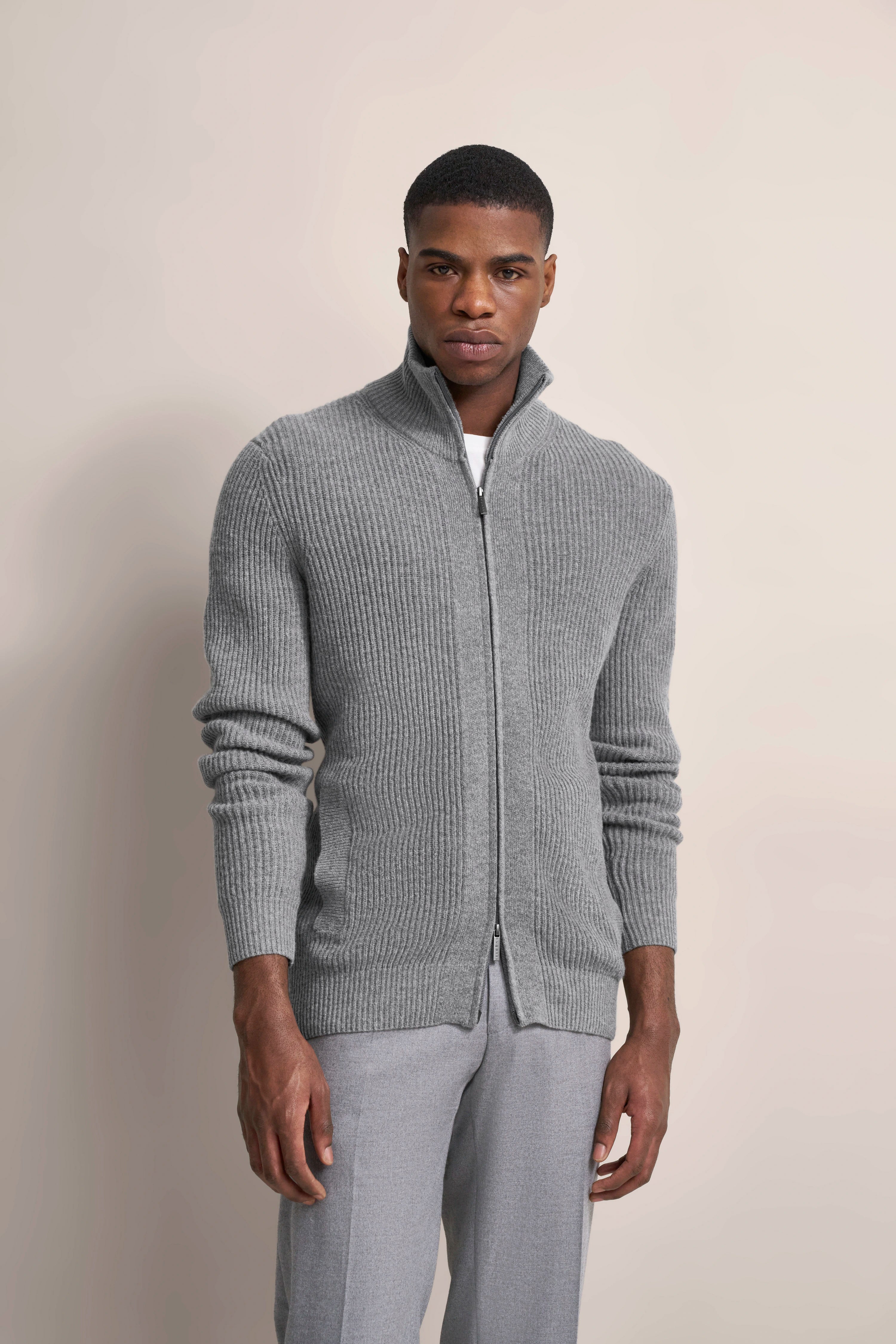Grauer Strick-Cardigan mit Reißverschluss, graue Hose, Model stehend, frontal, neutraler Hintergrund.