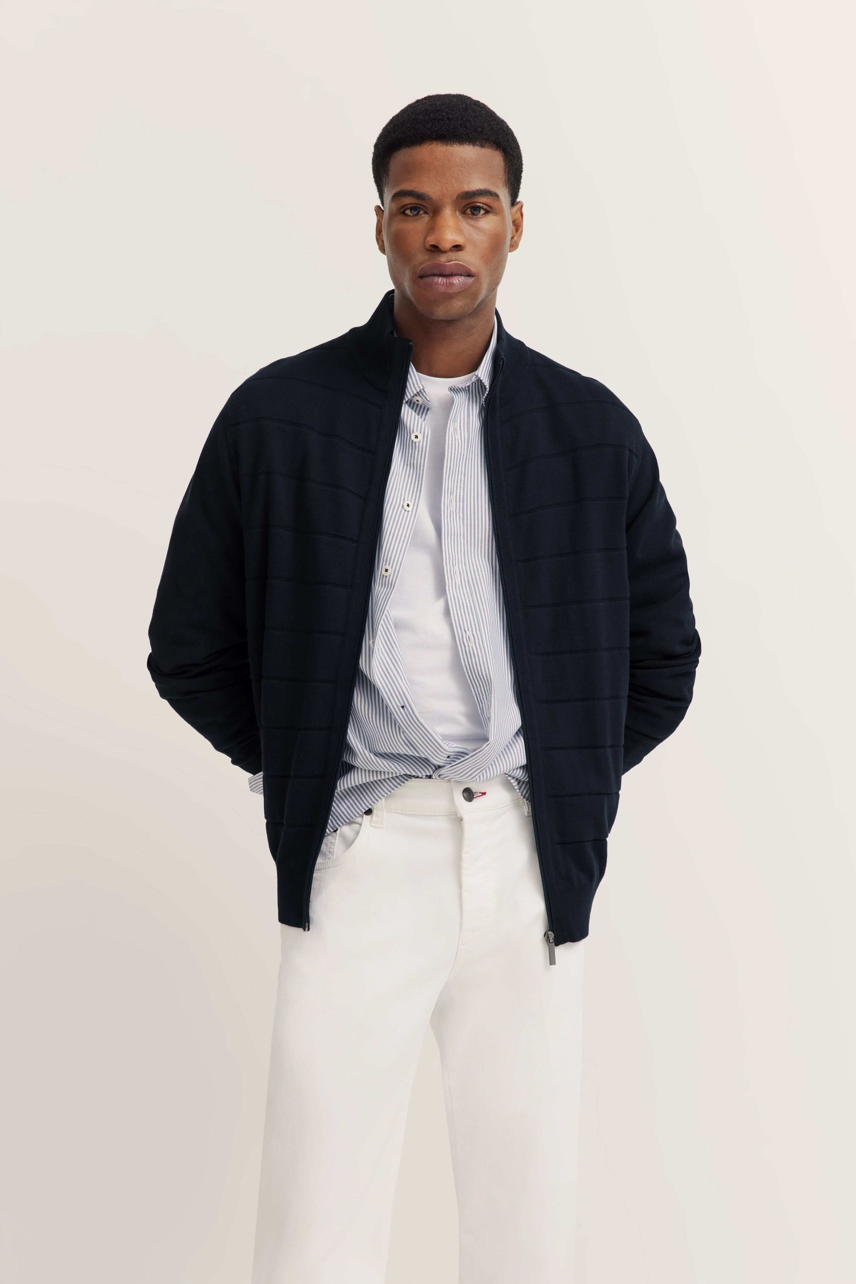bugatti modelbeeld: Man in zwarte jas, gestreept overhemd, wit shirt en witte broek, handen in zakken.