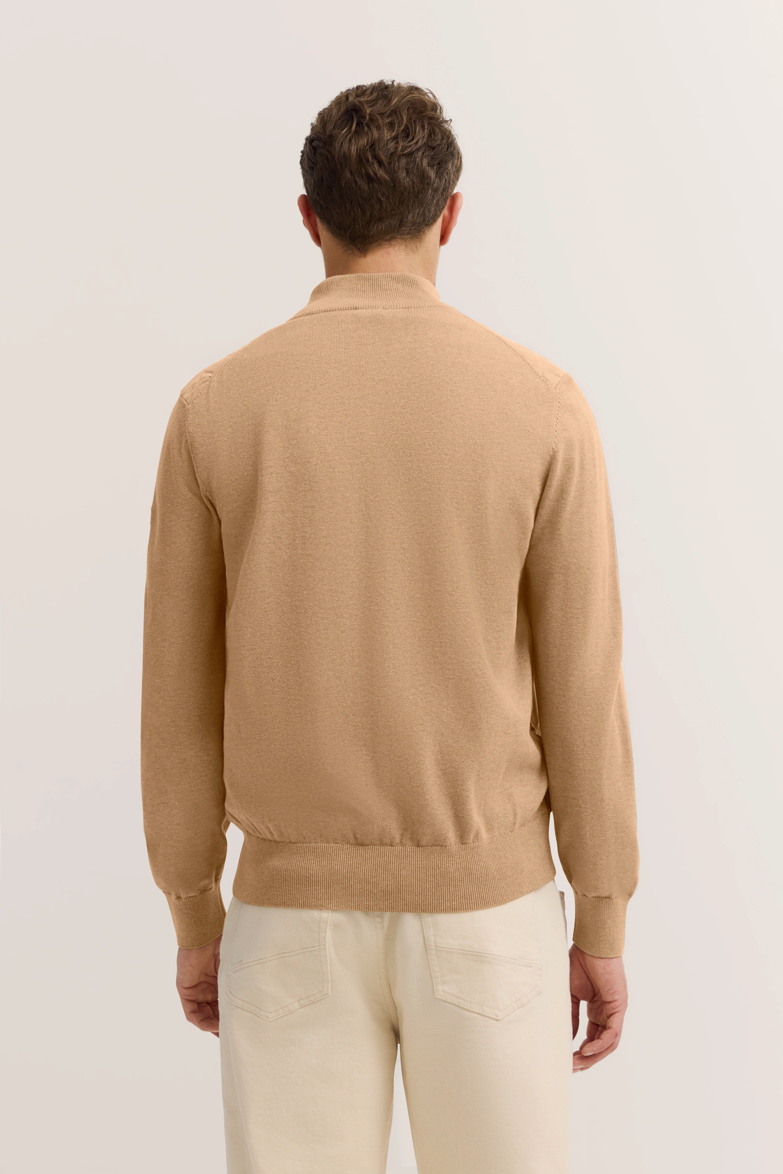 Achteraanzicht model in beige bugatti trui en crèmekleurige broek, ontspannen pose.