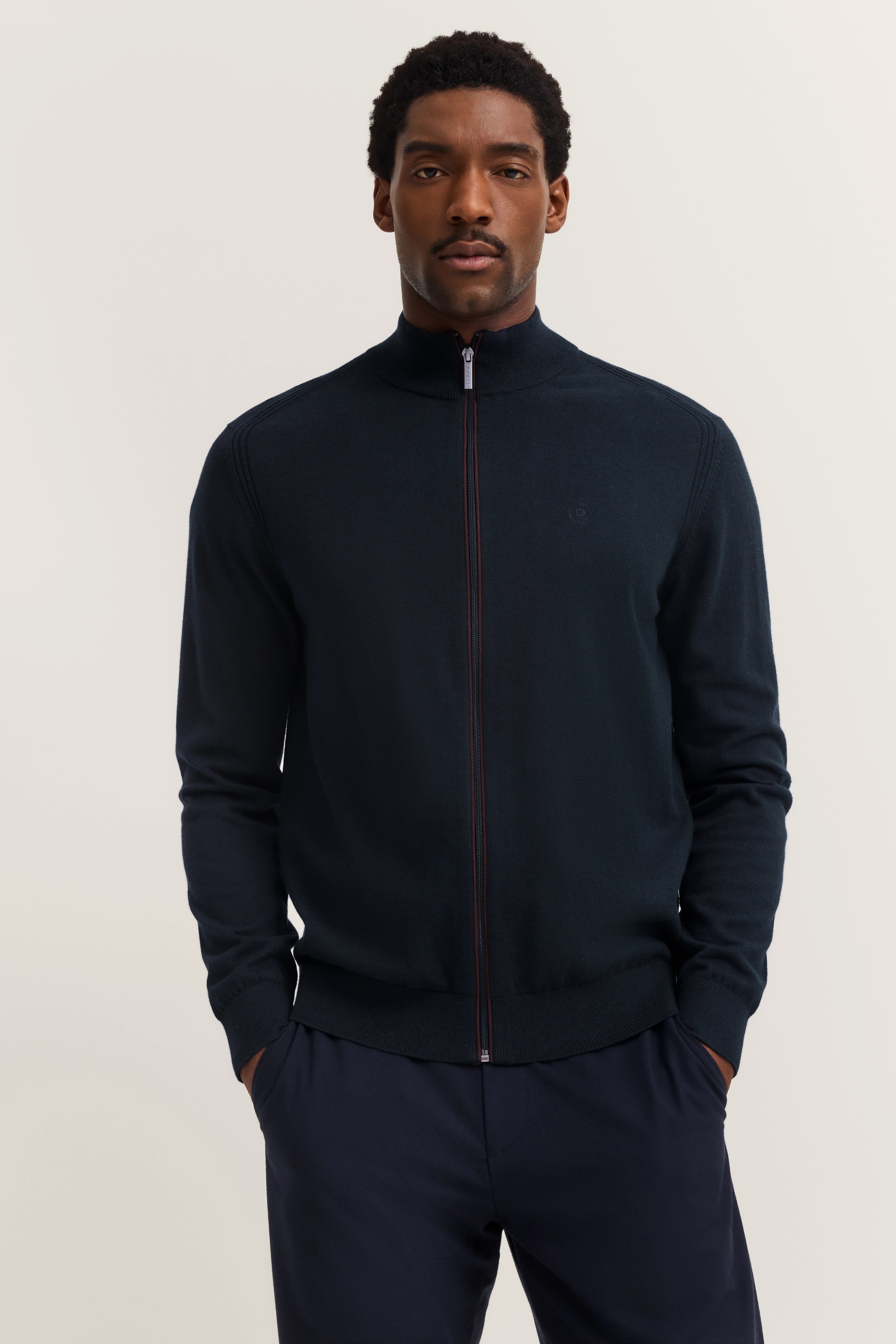 Modelbild mit schwarzem bugatti Zip-Pullover und dunkelblauer Hose, Hände in Taschen, entspannte Pose.