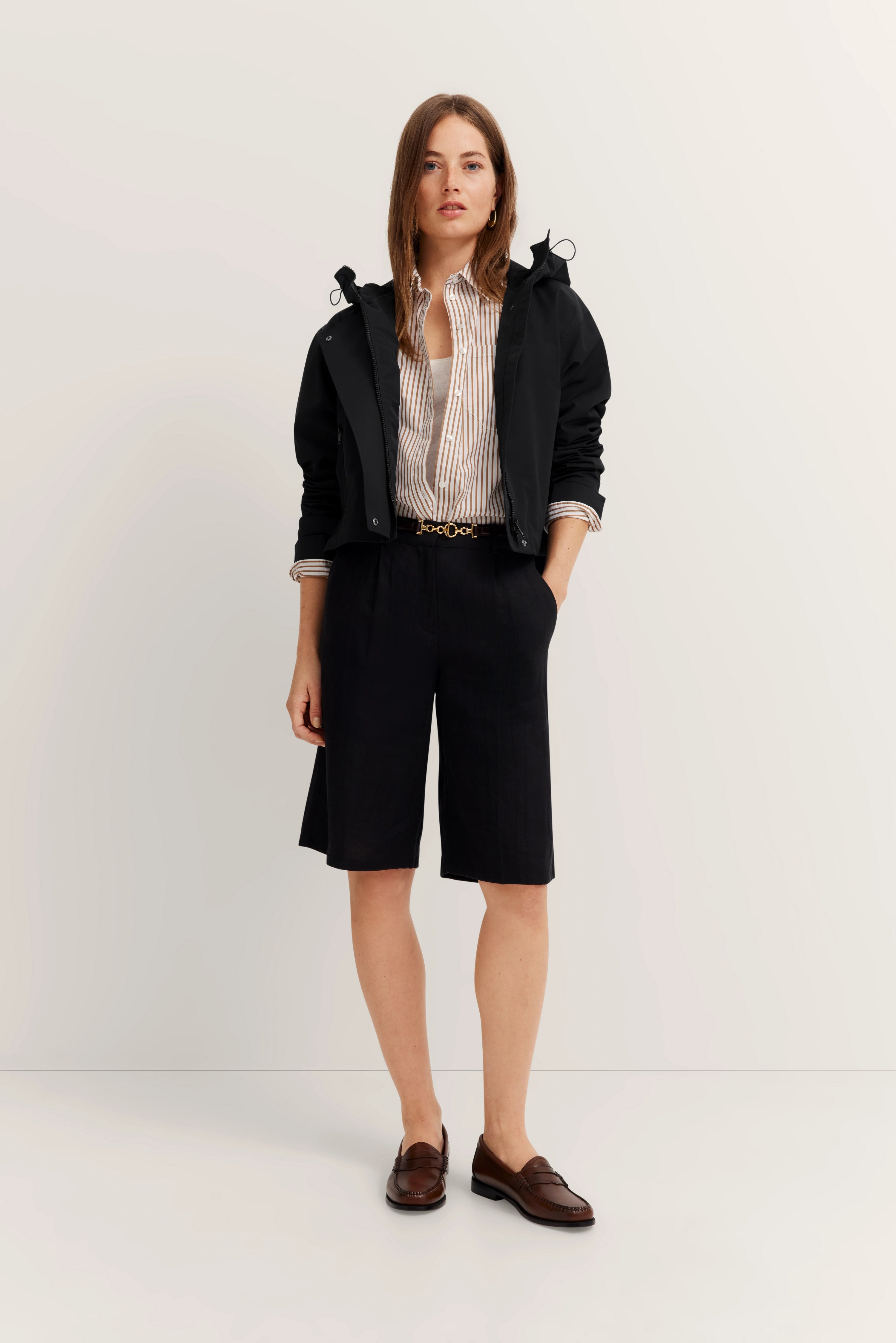 Model in zwarte jas, gestreept overhemd, zwarte shorts, bruine loafers