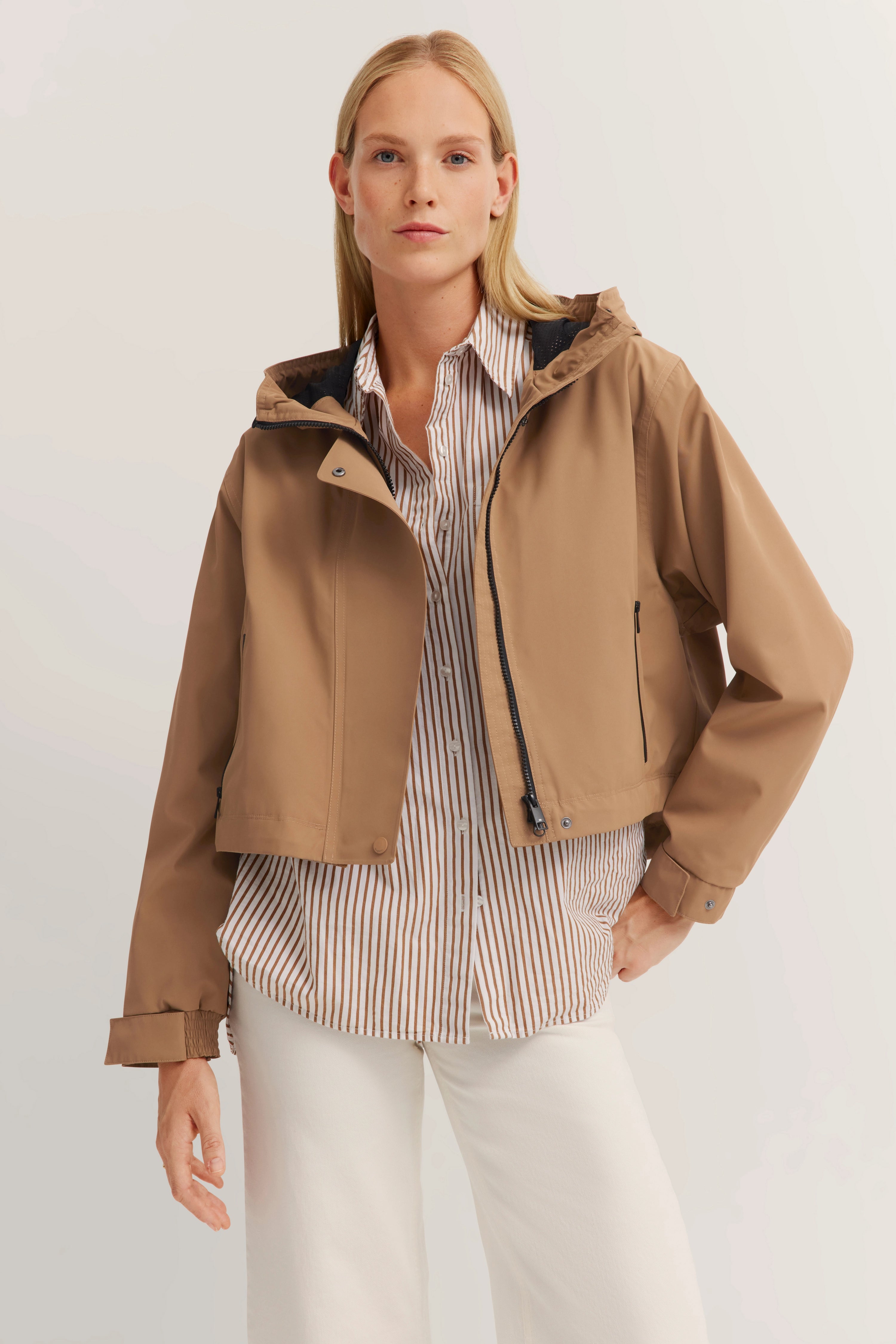 bugatti-model draagt beige jas, gestreept hemd en witte broek in ontspannen pose.