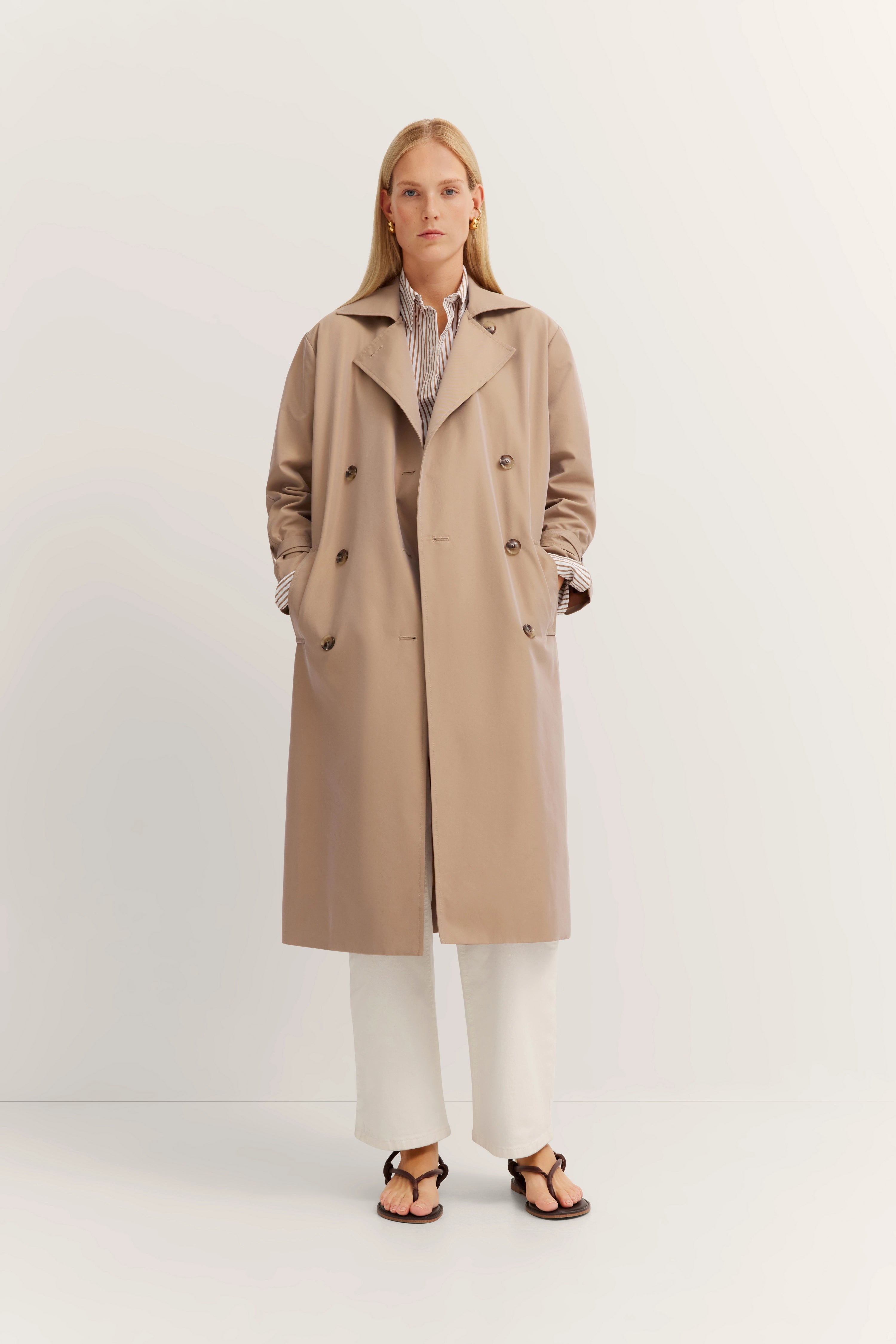 Beige bugatti trenchcoat met gestreept overhemd en witte broek, model in ontspannen pose.