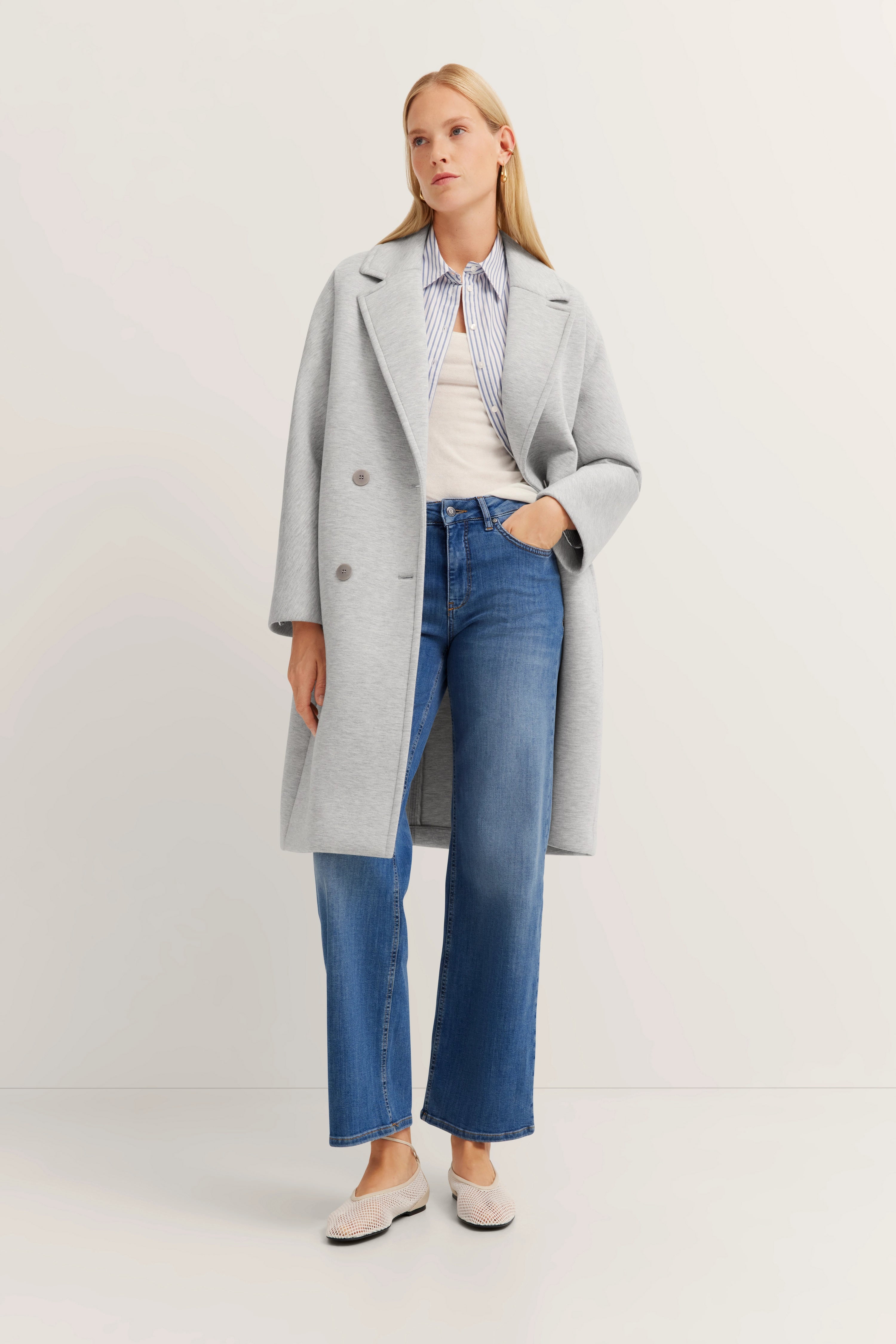 Grauer Mantel, gestreiftes Hemd, weiße Bluse, blaue Jeans, Model steht entspannt, bugatti Stil.