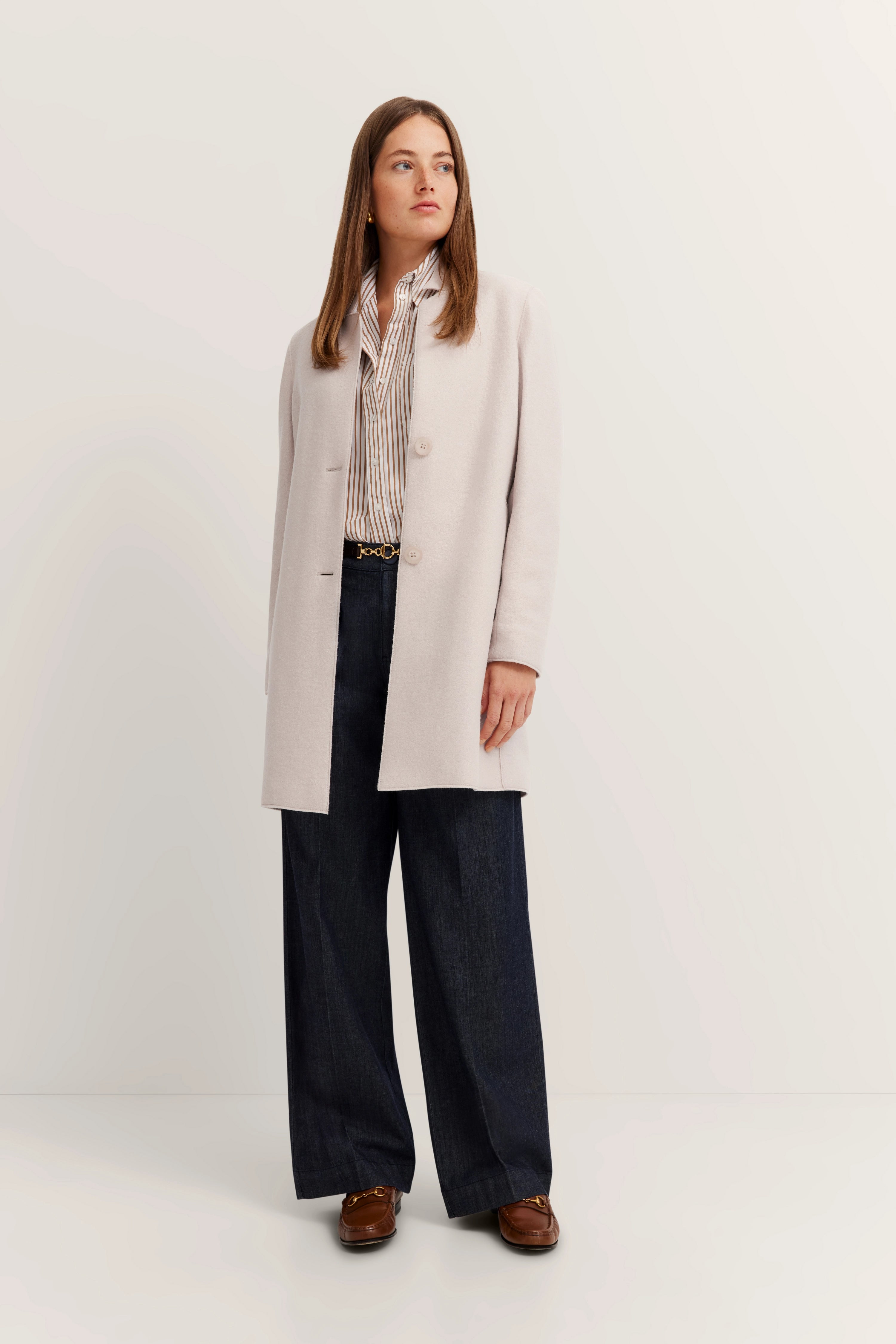 Model: Vrouw in crèmekleurige mantel, gestreepte blouse, blauwe wijde broek, bruine schoenen, ontspannen houding.