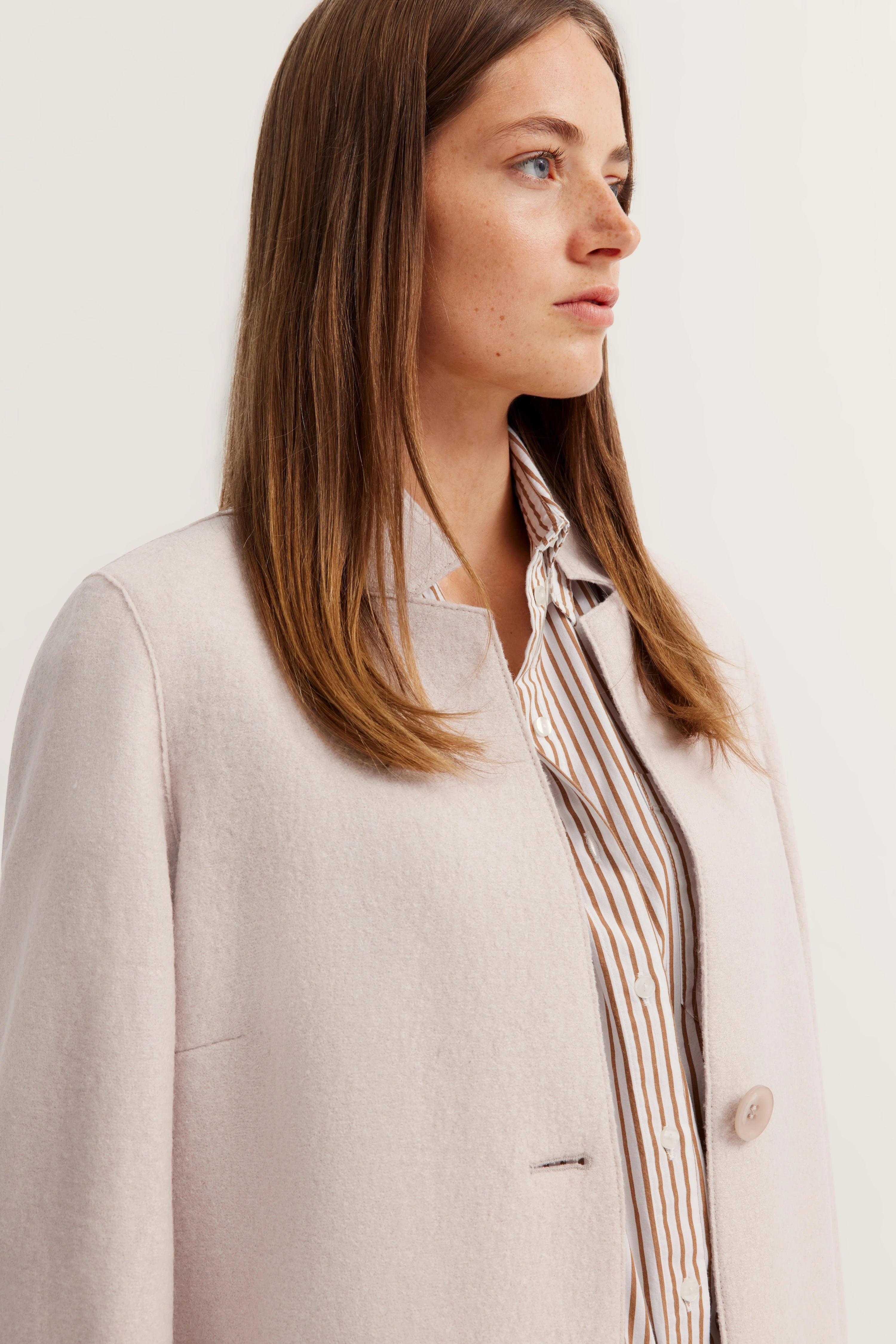Model in crèmekleurige bugatti-jas over gestreepte blouse, zijportret.
