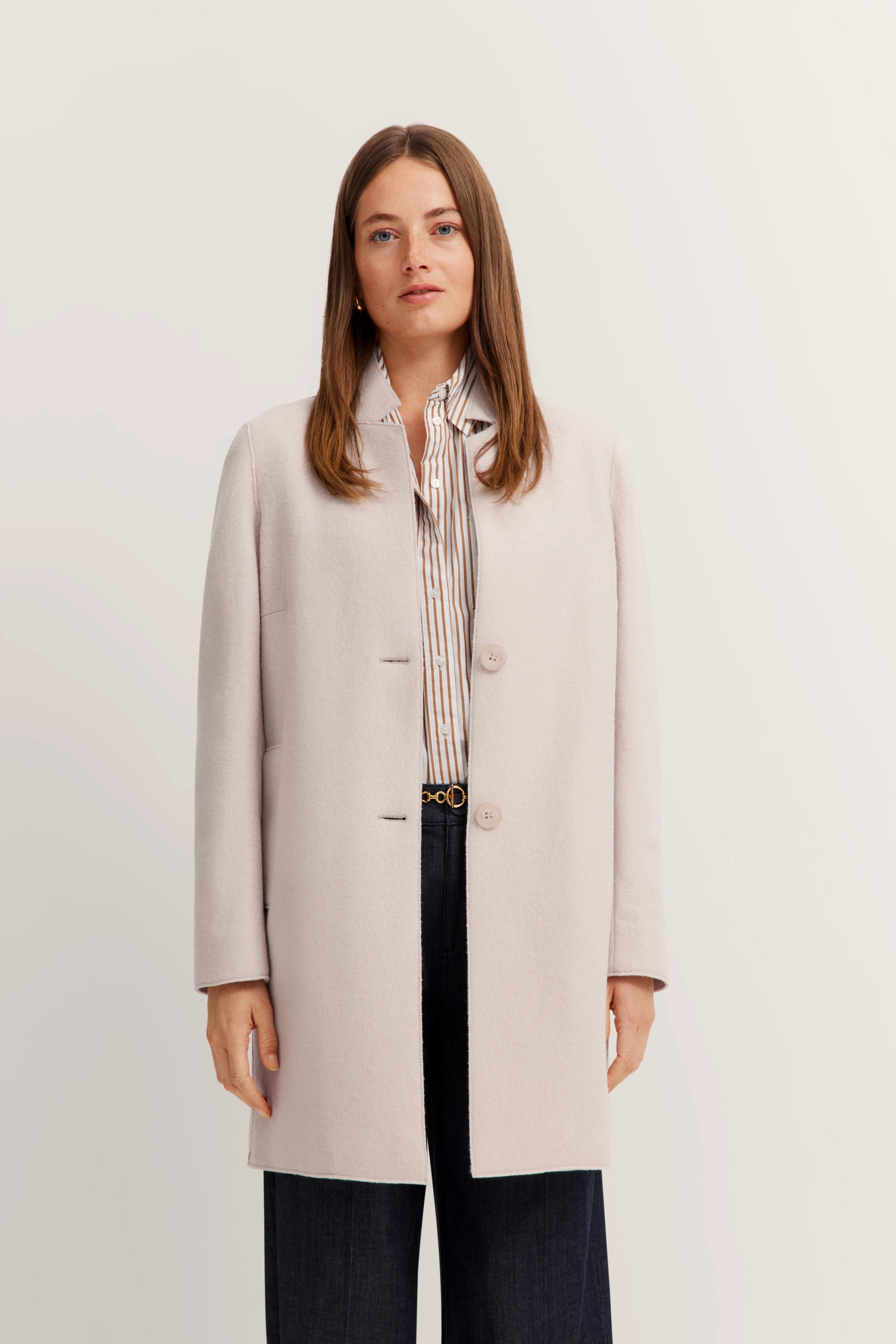 Model draagt lichte mantel, gestreepte blouse, donkere jeans. Elegant poseren, frontaal.