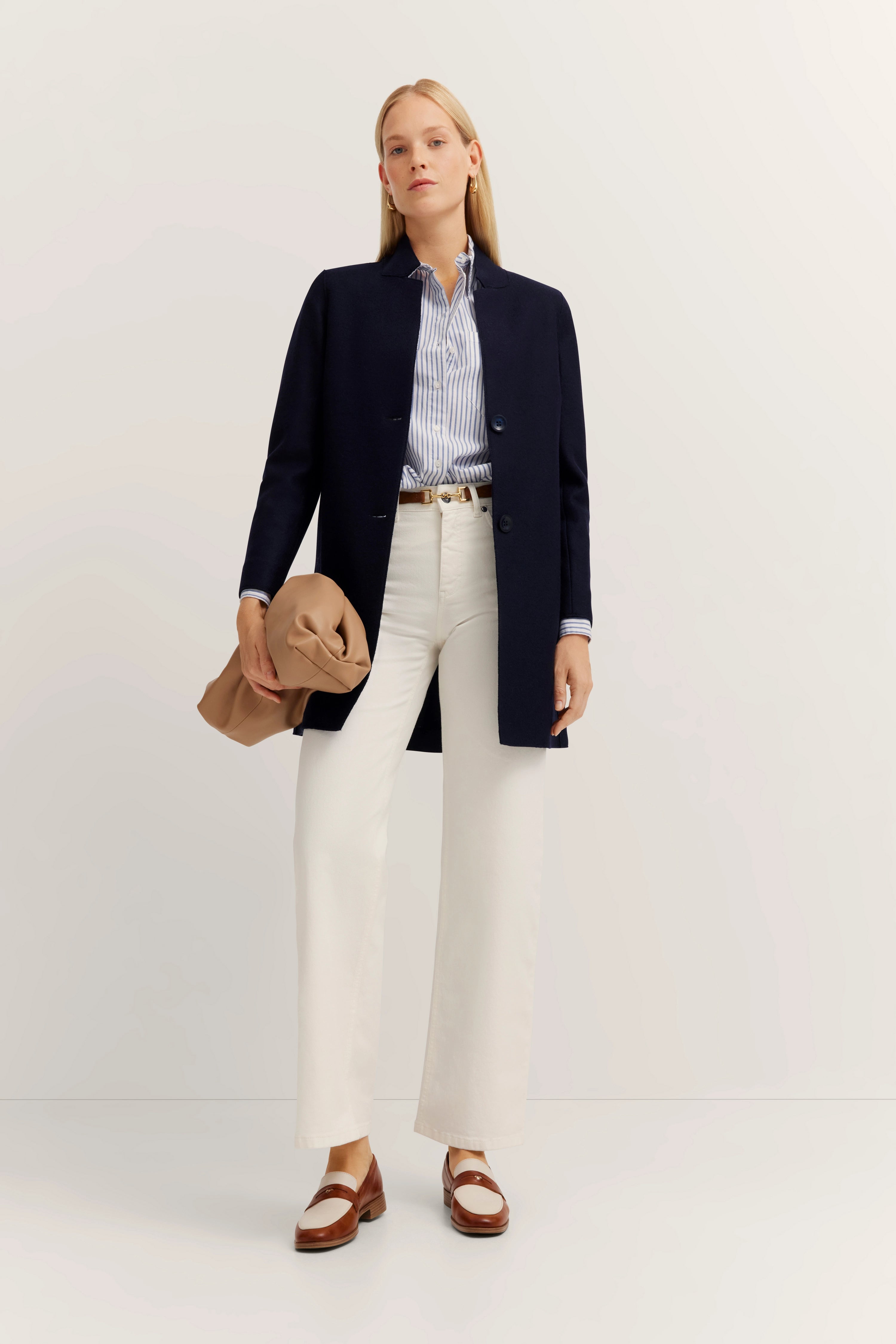 Elegant model in een marineblauwe jas, gestreept overhemd en witte broek. Houdt een beige tas vast.