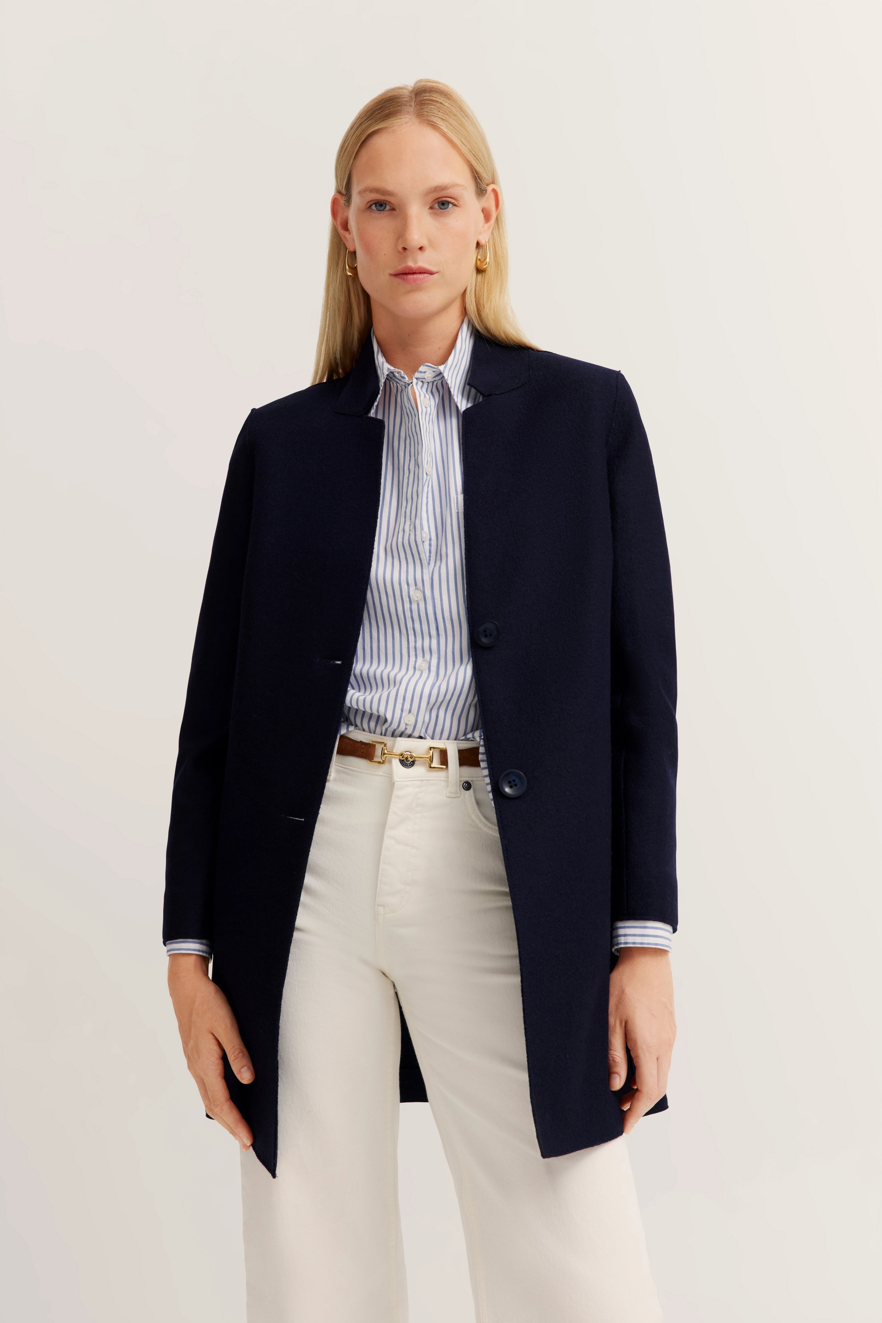 Man in marineblauwe jas, gestreept overhemd en witte broek, poseert ontspannen.