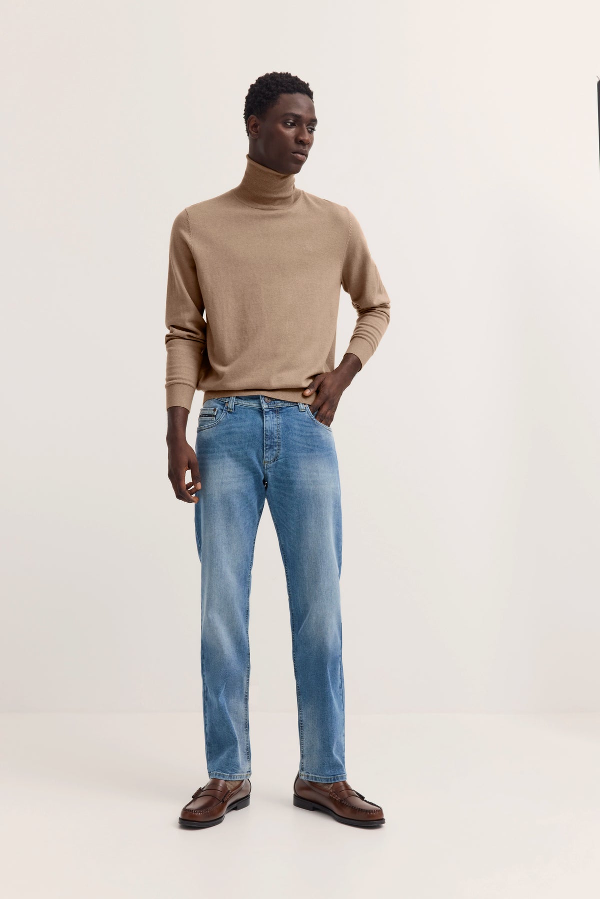 Model in beigem Rollkragenpullover, blauer Jeans und braunen Schuhen, stehend mit Hand in Hüfte.
