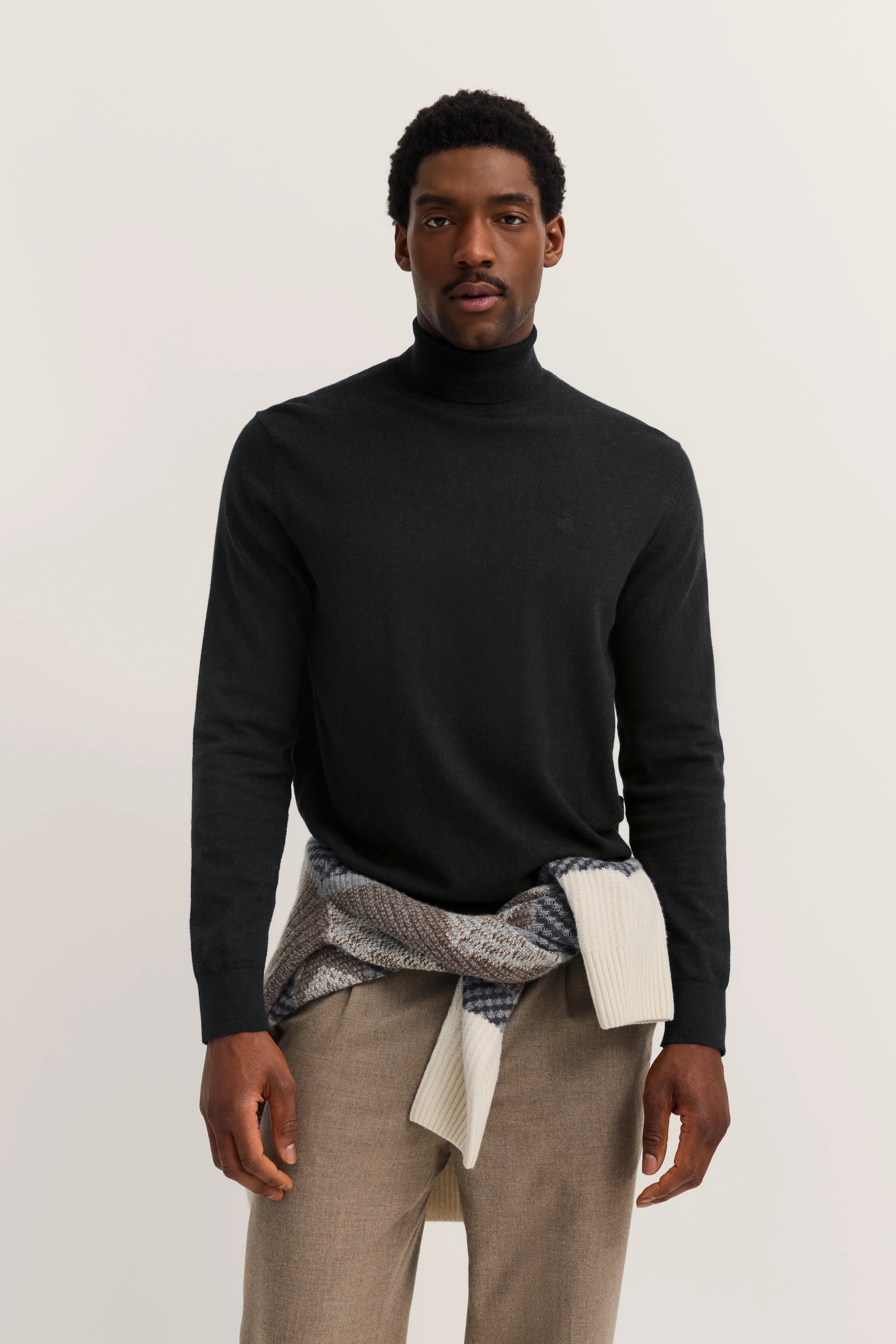 Schwarzer Rollkragenpullover, beigefarbene Hose, Strickpullover um die Taille, Modelbild stehend.