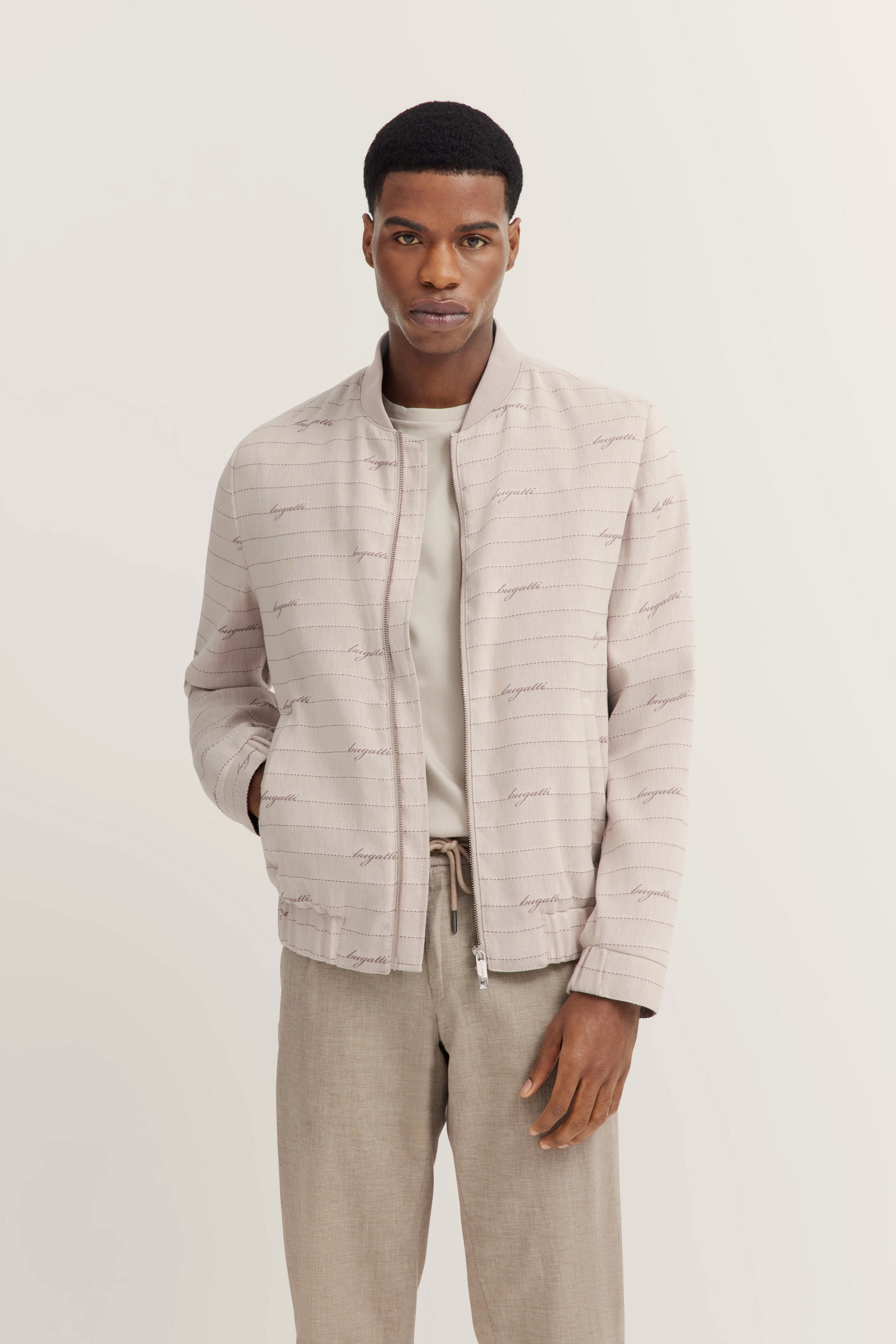 Model met beige bugatti-jas, T-shirt en broek. Ontspannen houding, handen in de zakken.