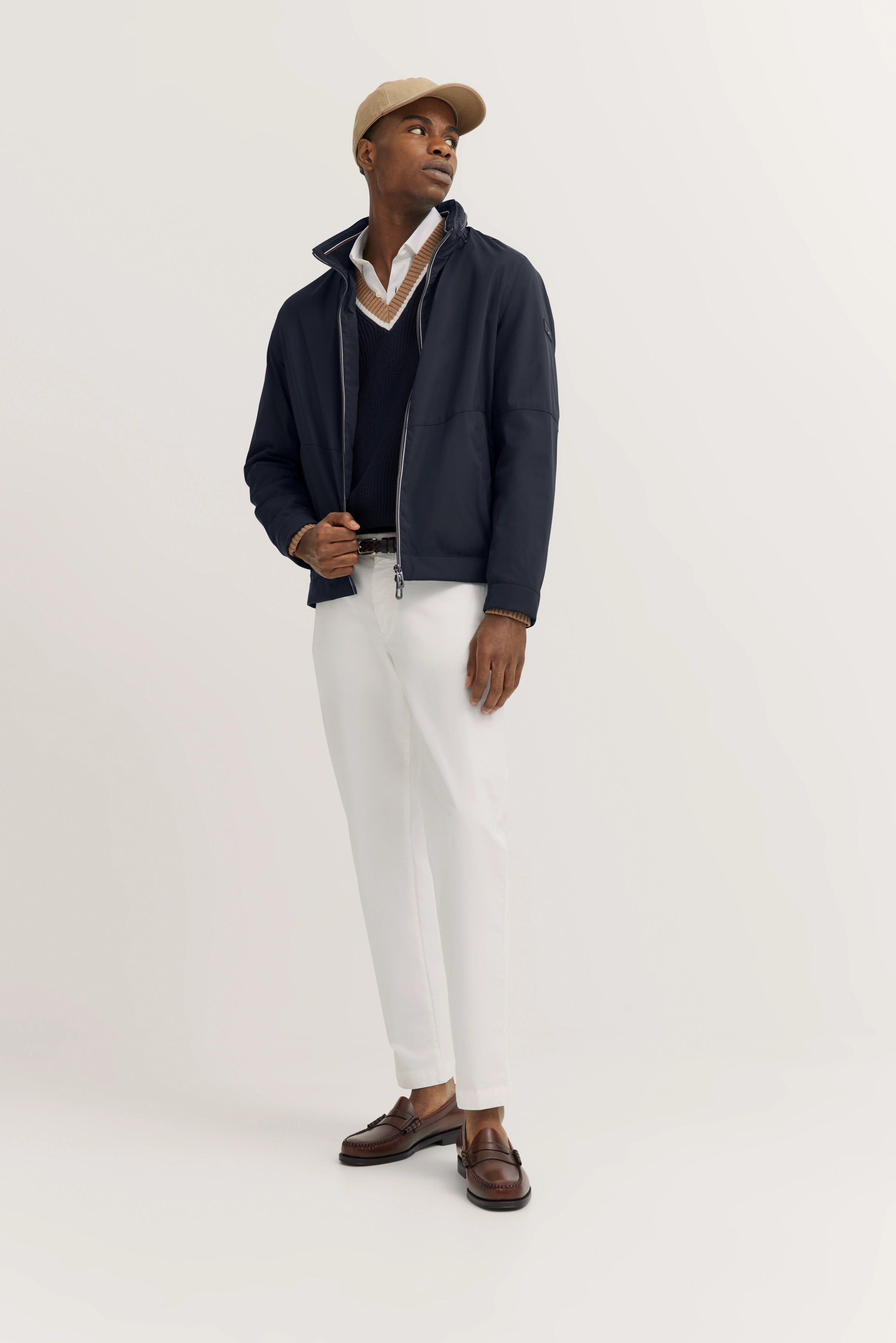 Navy Jacke, weißes Hemd, Pullover, beige Kappe, weiße Hose, braune Schuhe, Model stehend, Blick nach rechts.