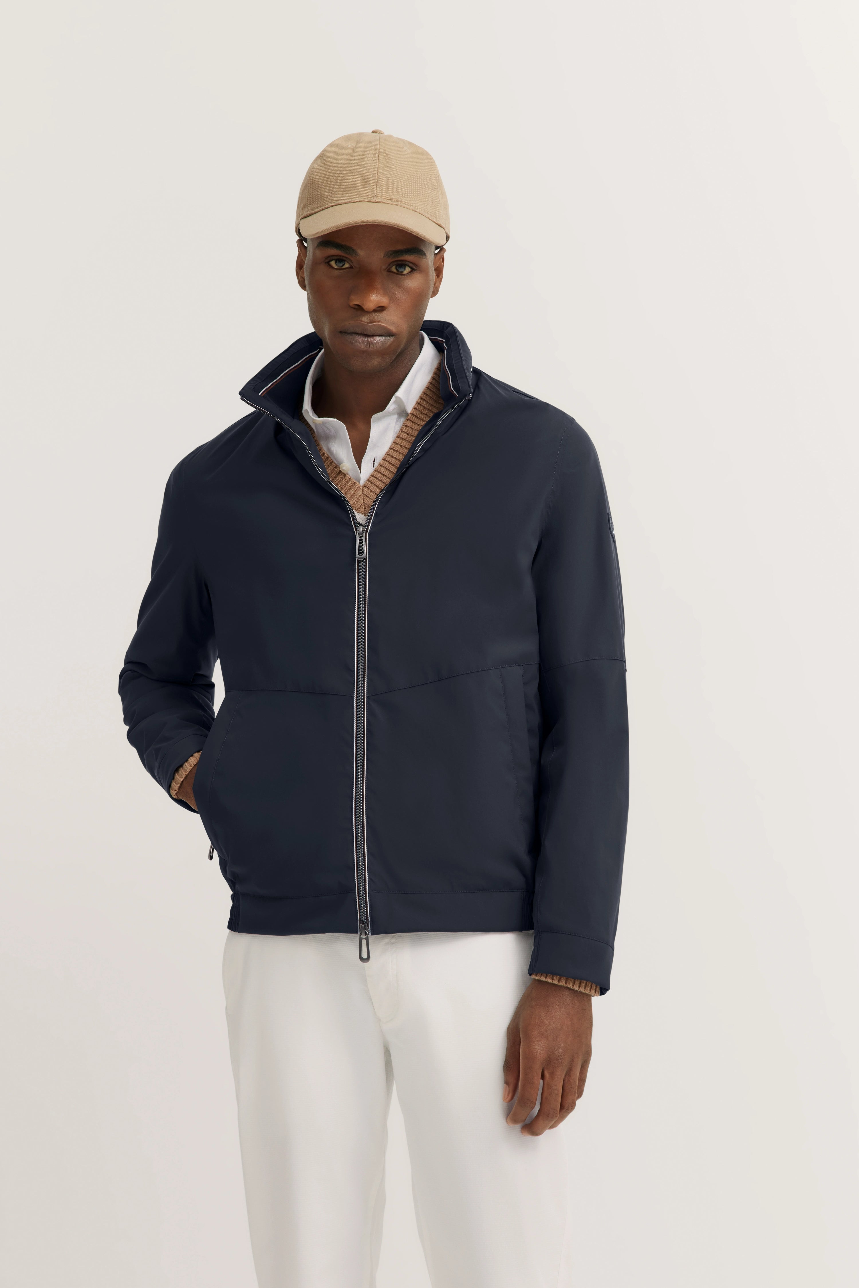 Model in marineblauwe bugatti jas, beige muts, wit overhemd, trui en witte broek. Ontspannen pose.