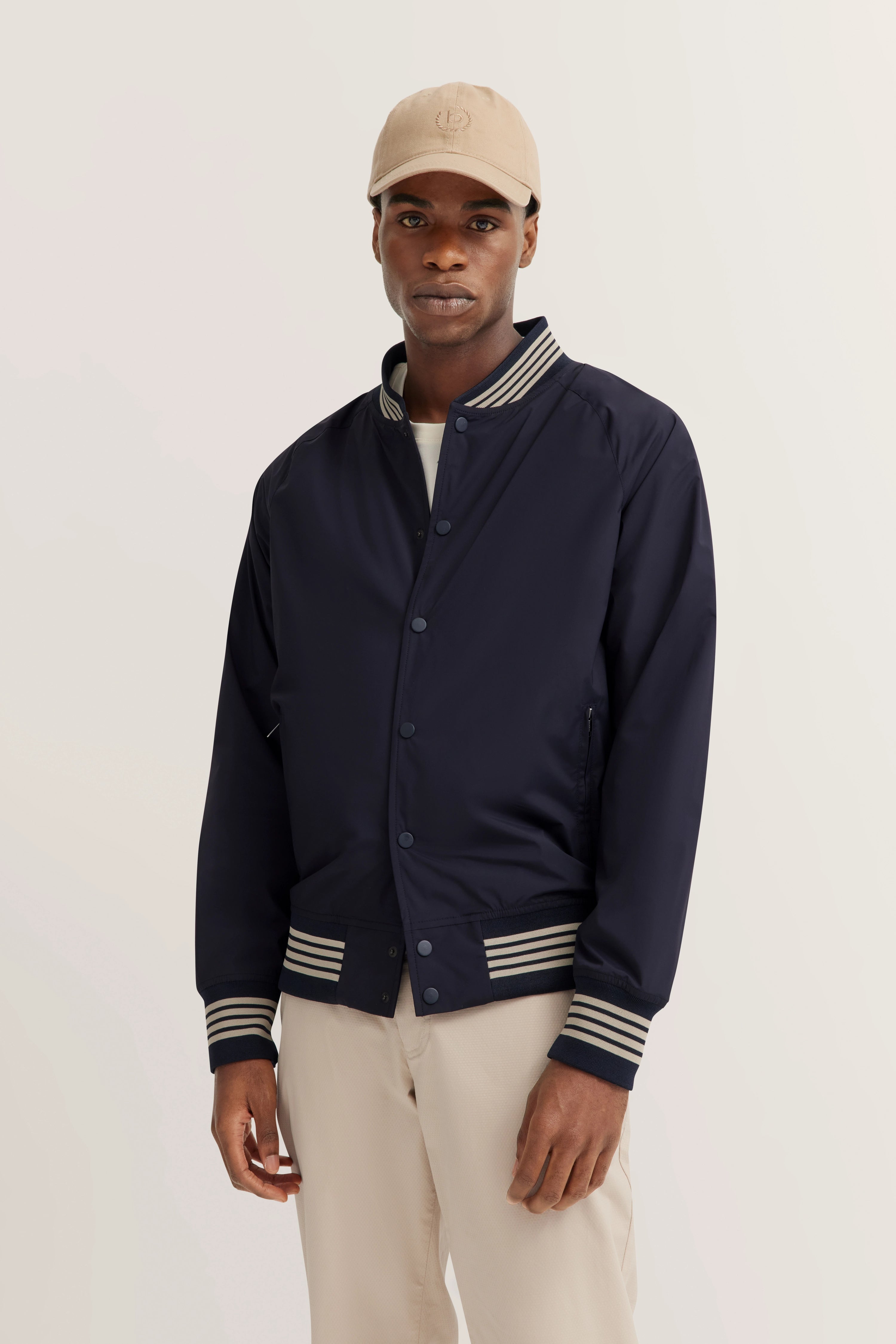 Marineblauw collegejack met beige pet, model staand, ontspannen uitstraling.
