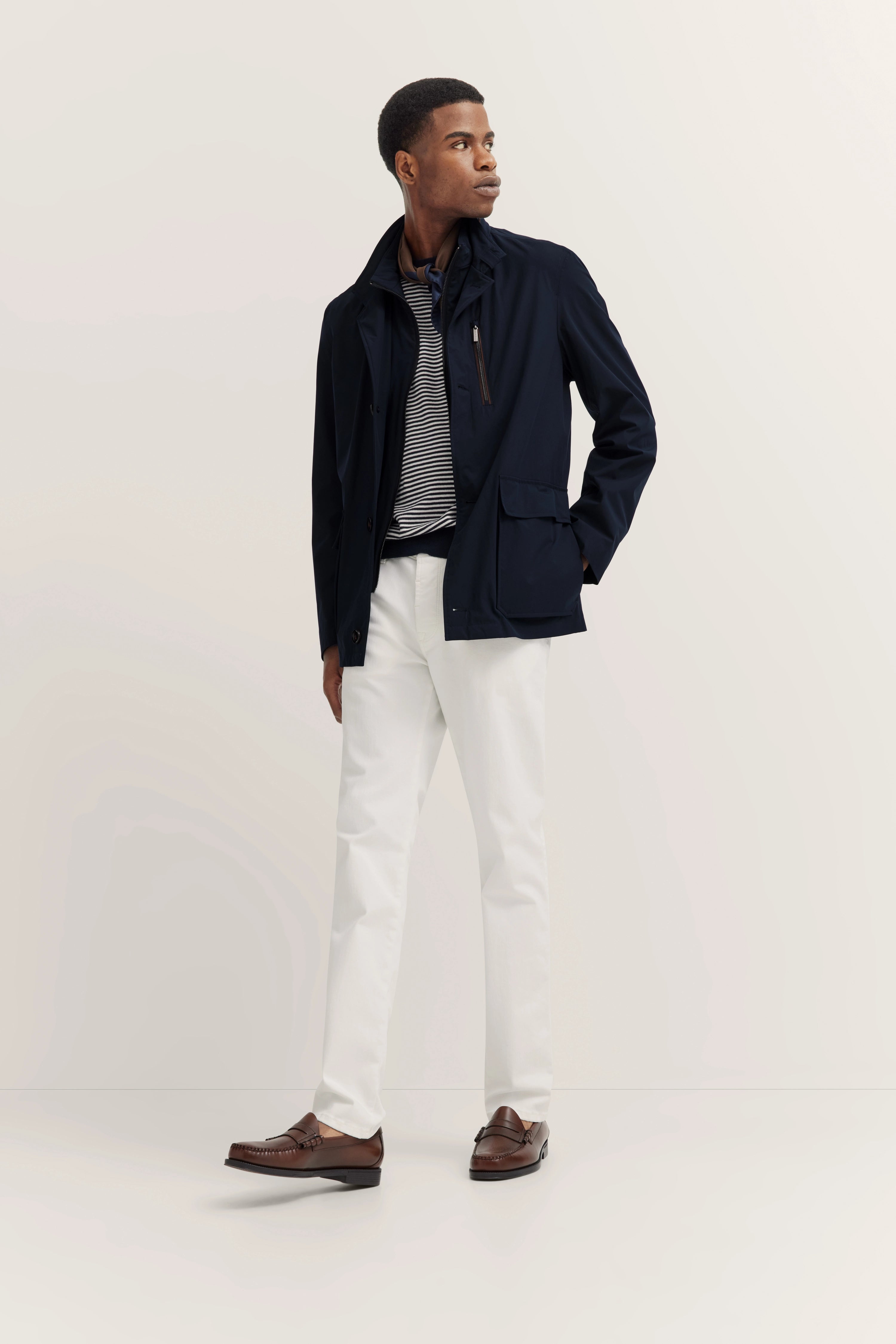 Model in donkerblauwe jas, gestreept shirt, witte broek. Nonchalant staand, kijkend opzij.