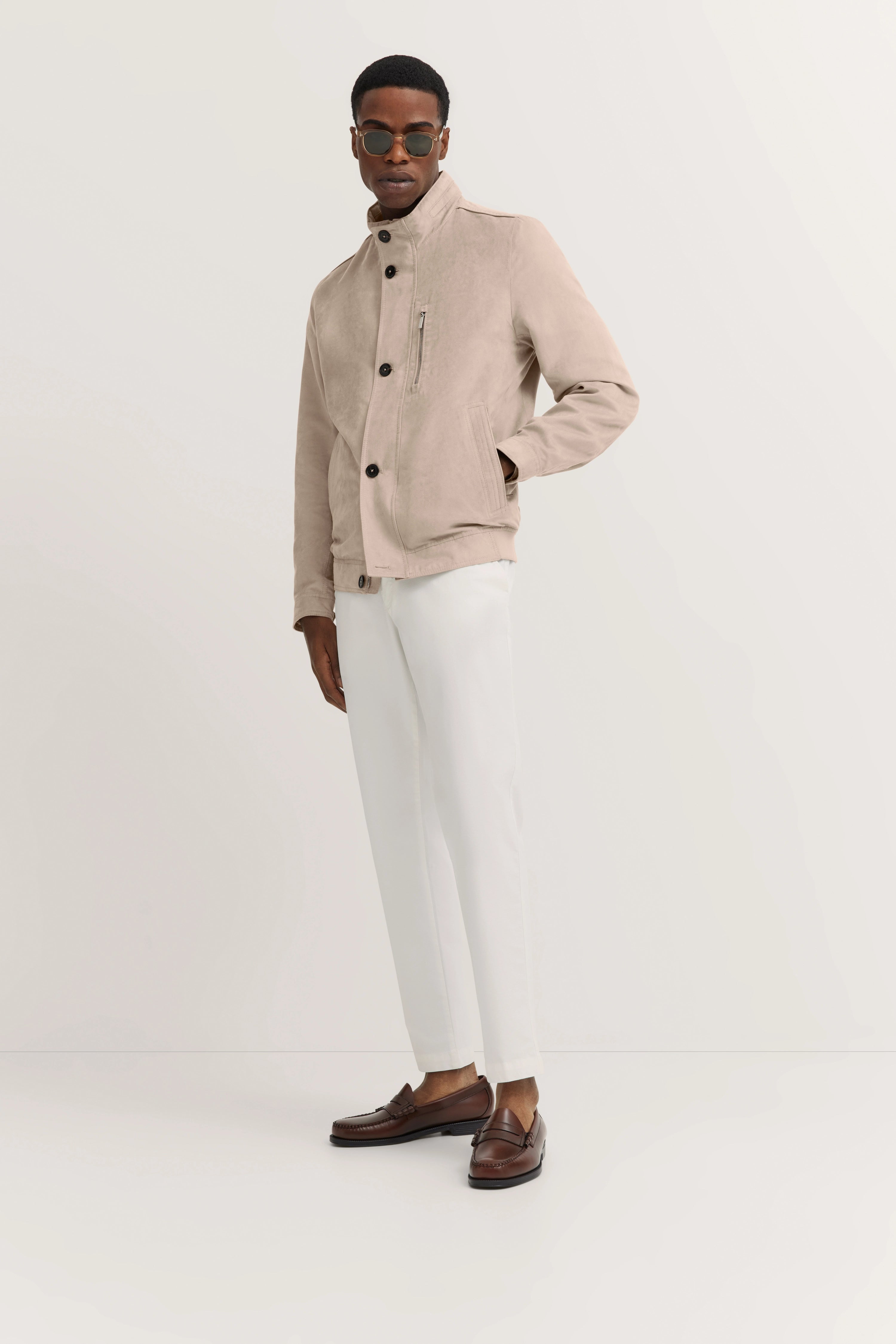 Beiger bugatti Blouson, weiße Hose, braune Loafer, legeres Modelbild, lässige Pose mit Händen in der Tasche.
