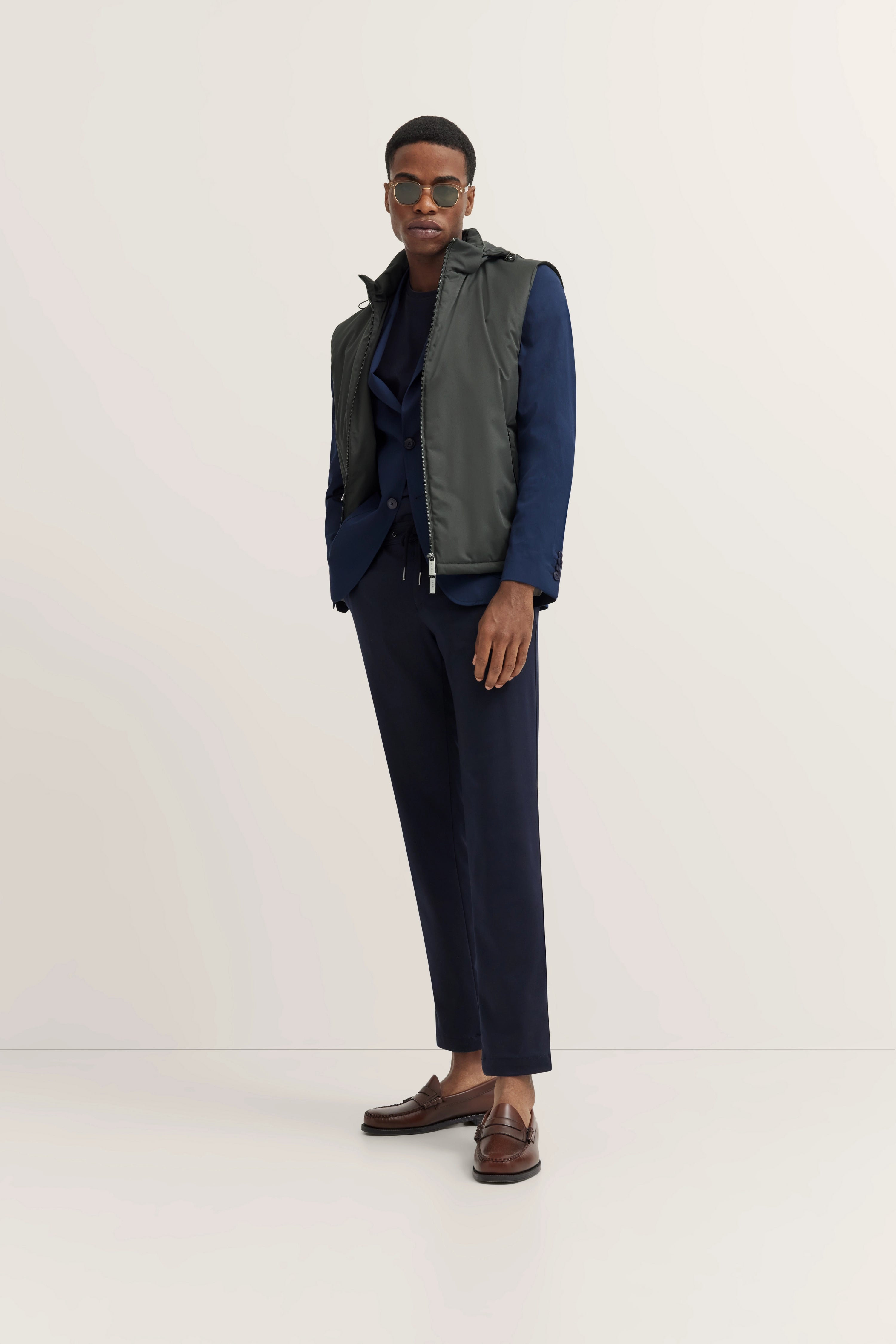 Model in marineblauw pak met grijs vest, bruine loafers. Ontspannen pose, handen in zakken.