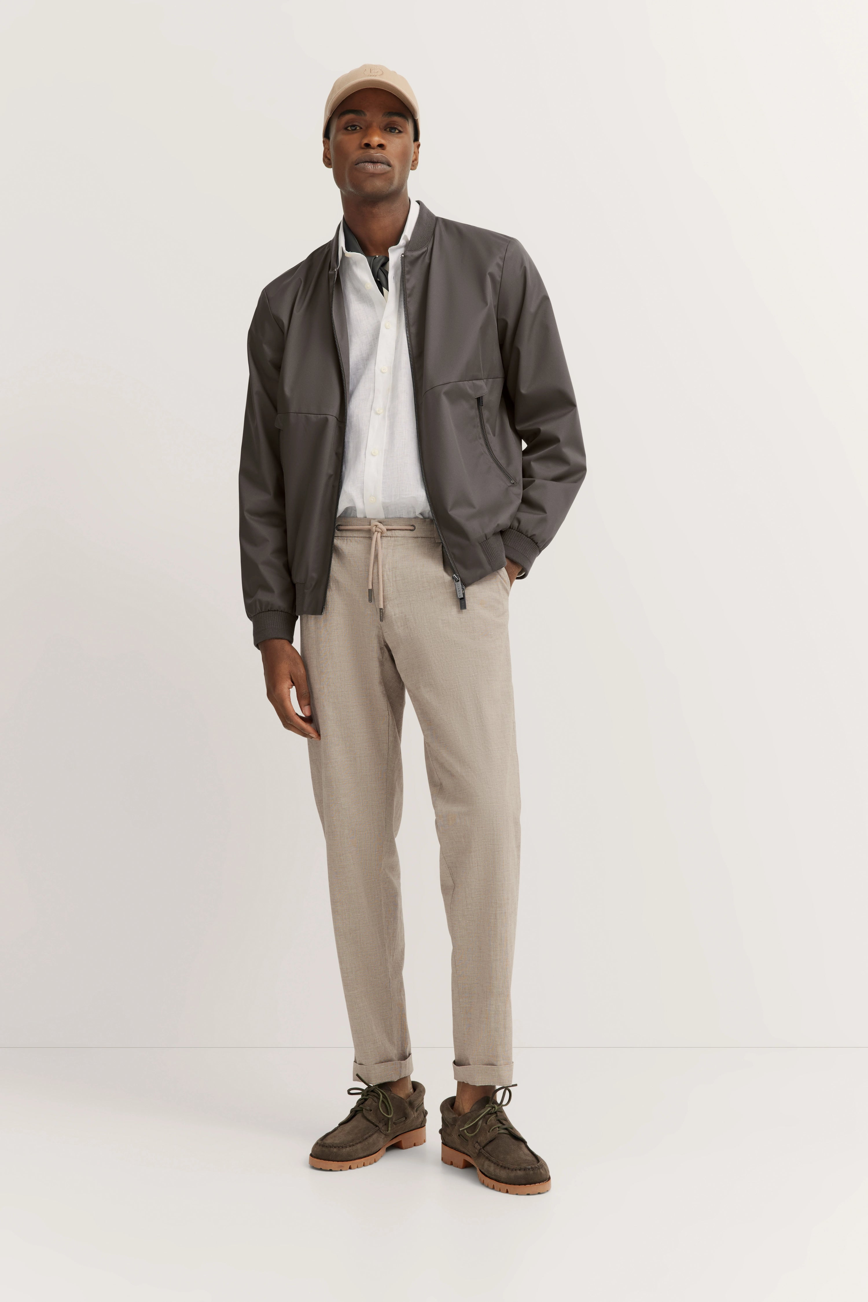 Model in grijze jas, wit overhemd en beige broek. Beige muts, bruine schoenen. Ontspannen houding.