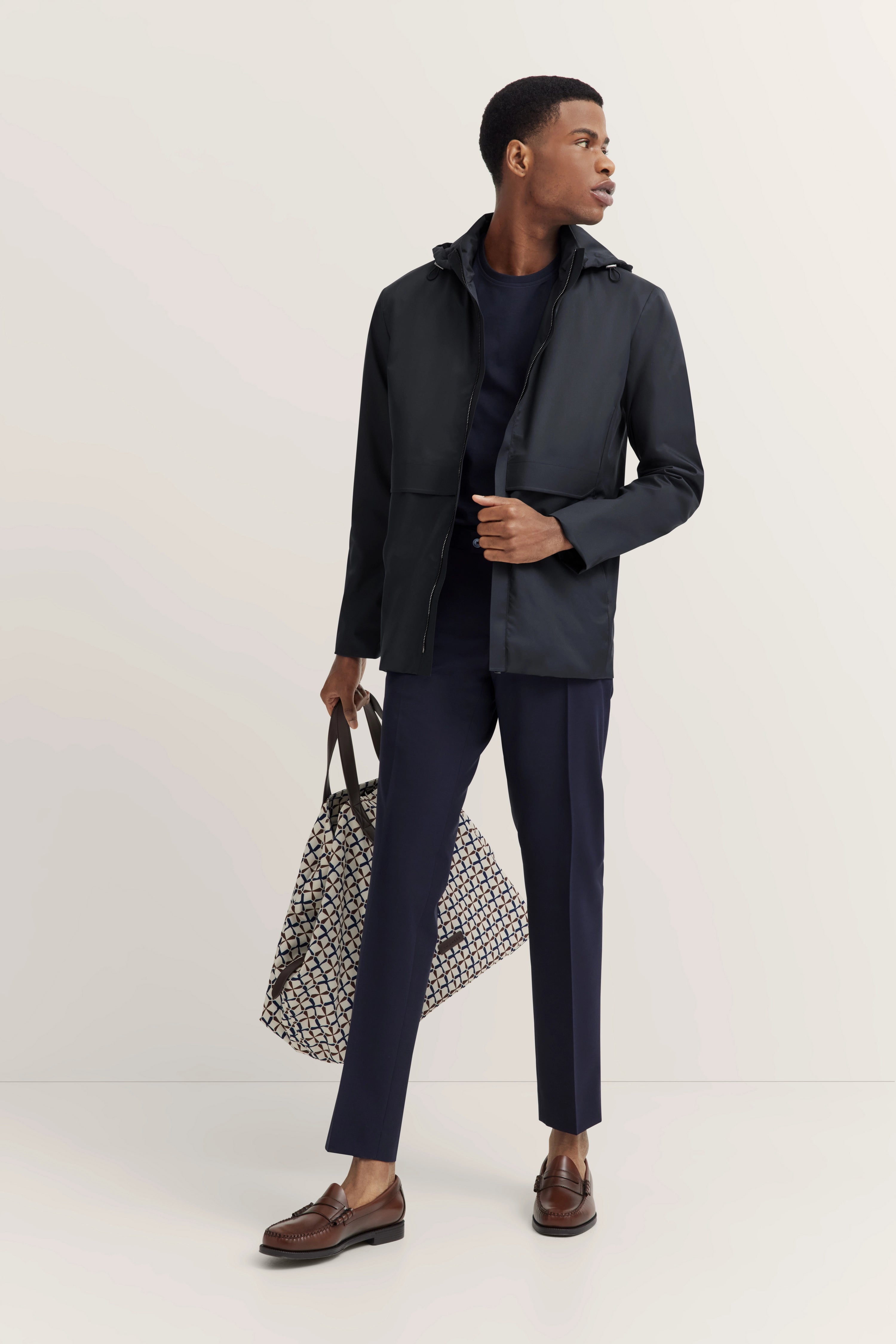 Model in donkerblauwe bugatti outfit, ontspannen poserend met een patroon tas en bruine loafers.