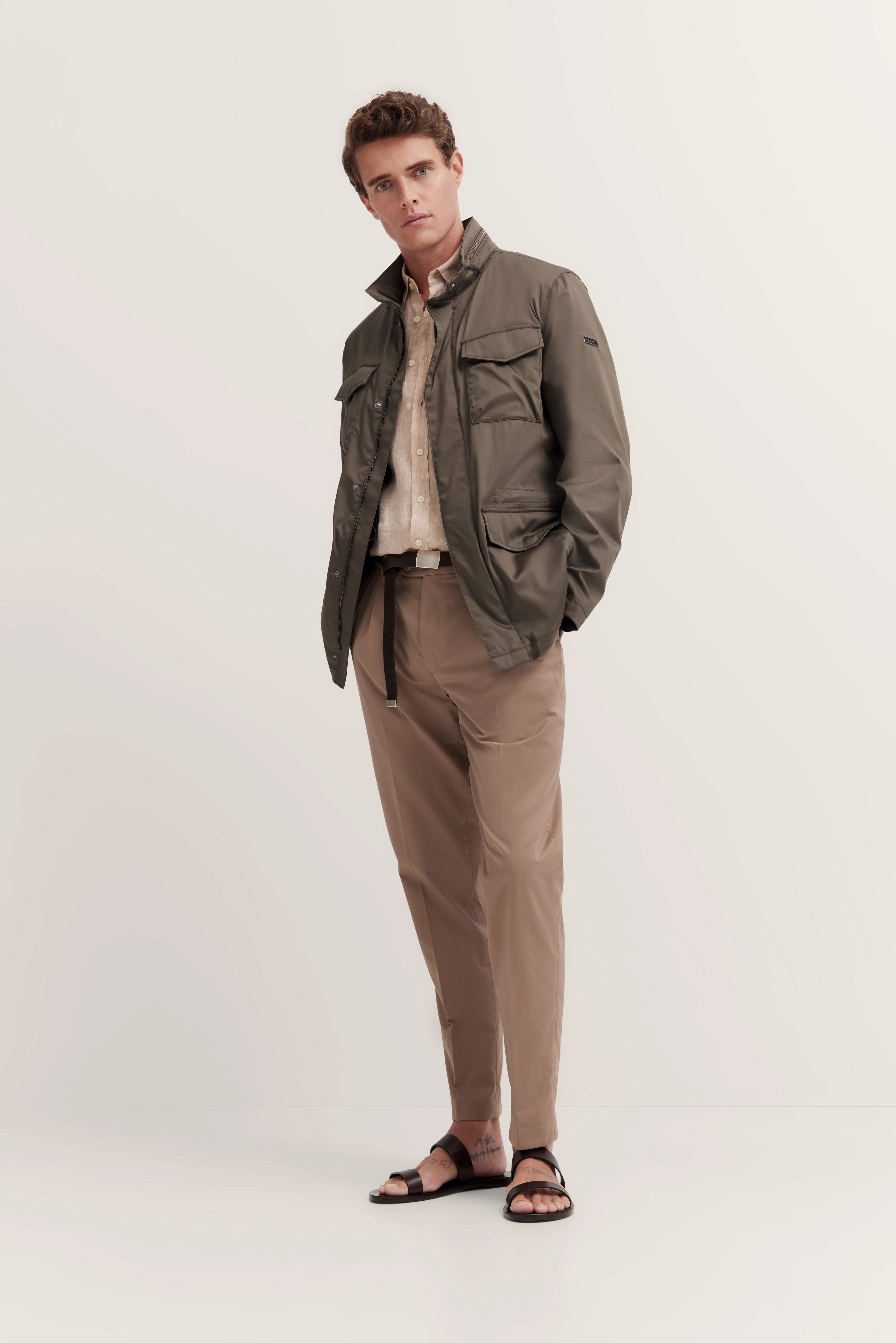 Model draagt een olijfgroene jack, beige overhemd, kaki broek, zwarte sandalen. Casual pose.
