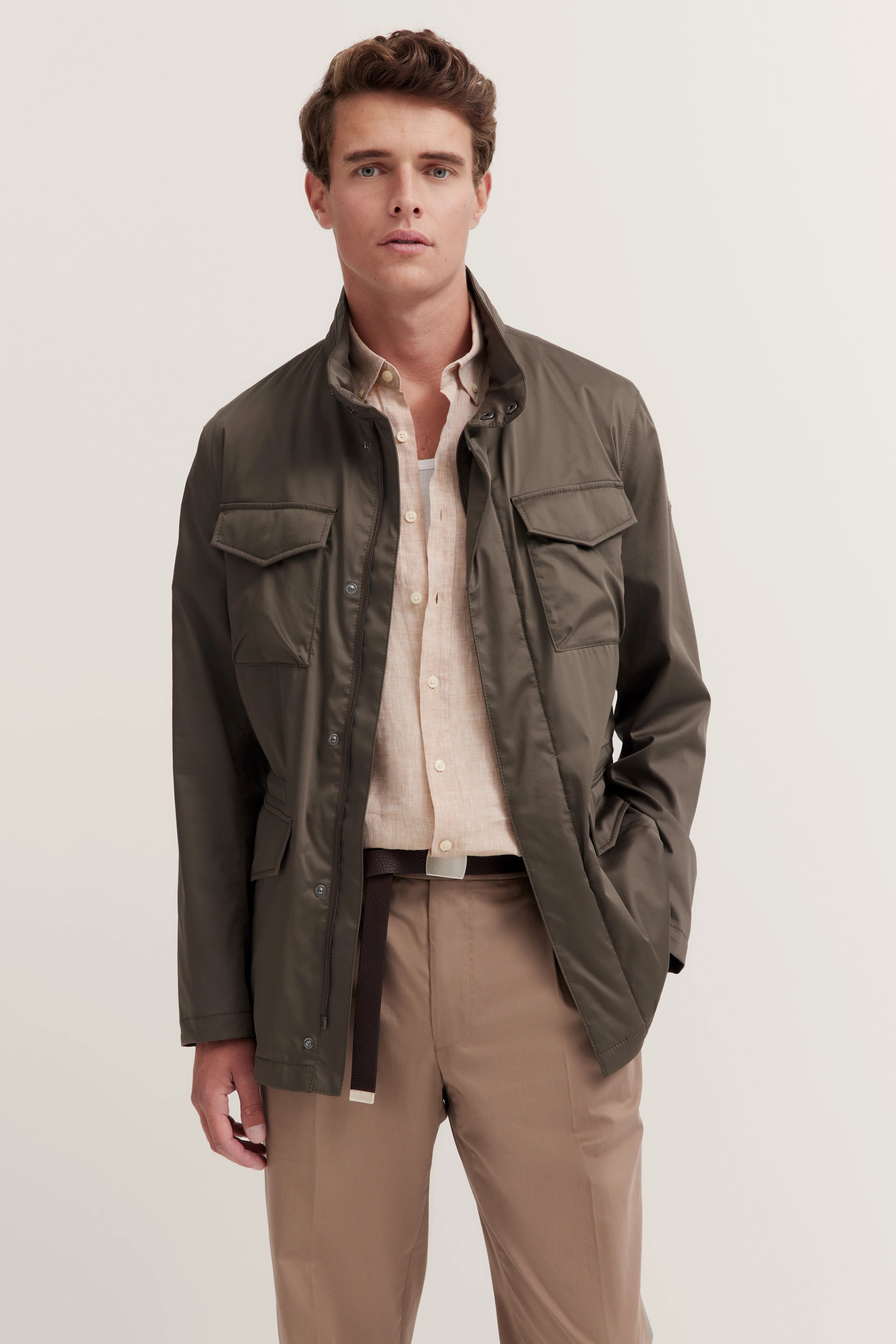 Model in olijfgroene bugatti jas, beige shirt en bruine broek, ontspannen pose met hand in zak.