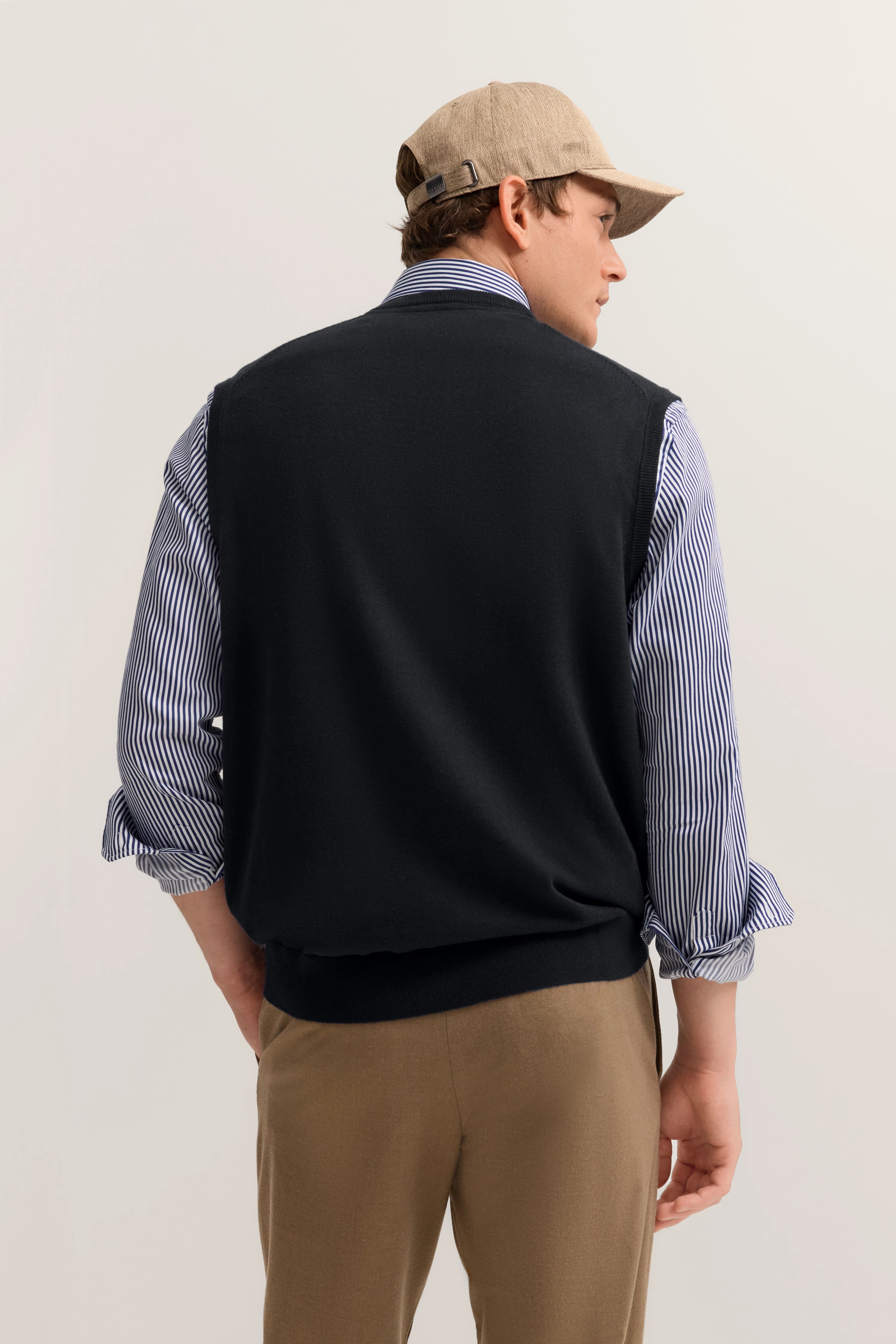 Bugatti Model, beige Hose, gestreiftes blaues Hemd, schwarzer Pullover, beige Cap, Rückansicht, legeres Posing.