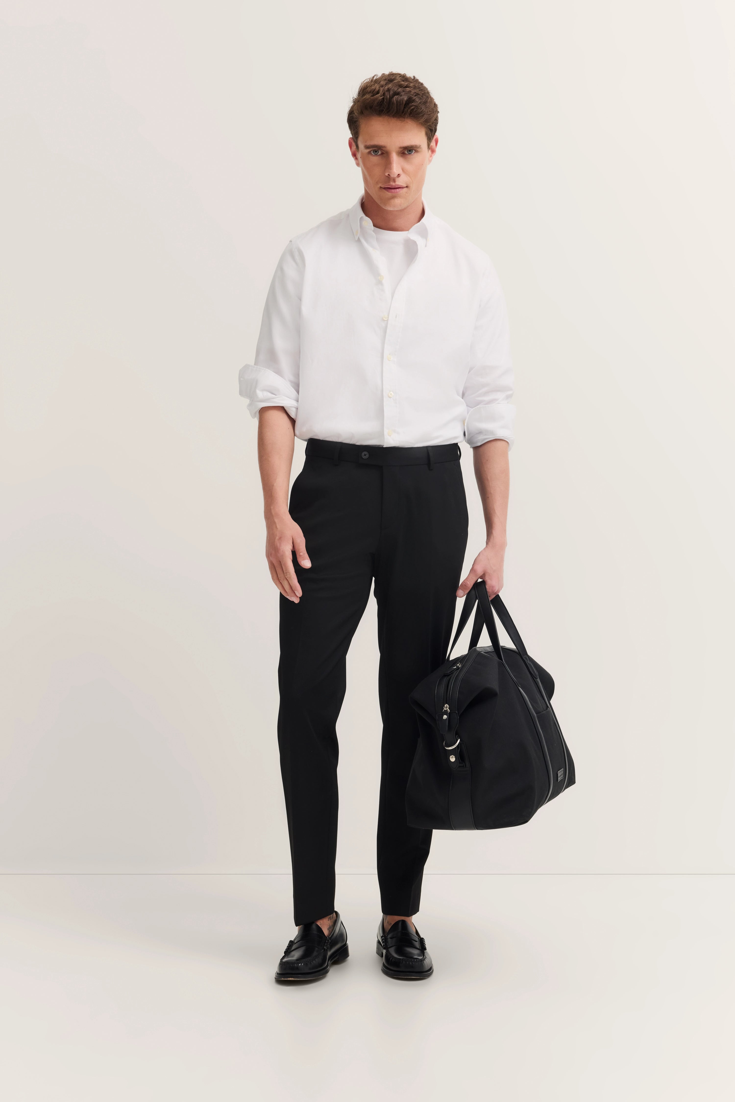 Model in white shirt, black pants, with black bag. Standing, relaxed hands, bugatti style.