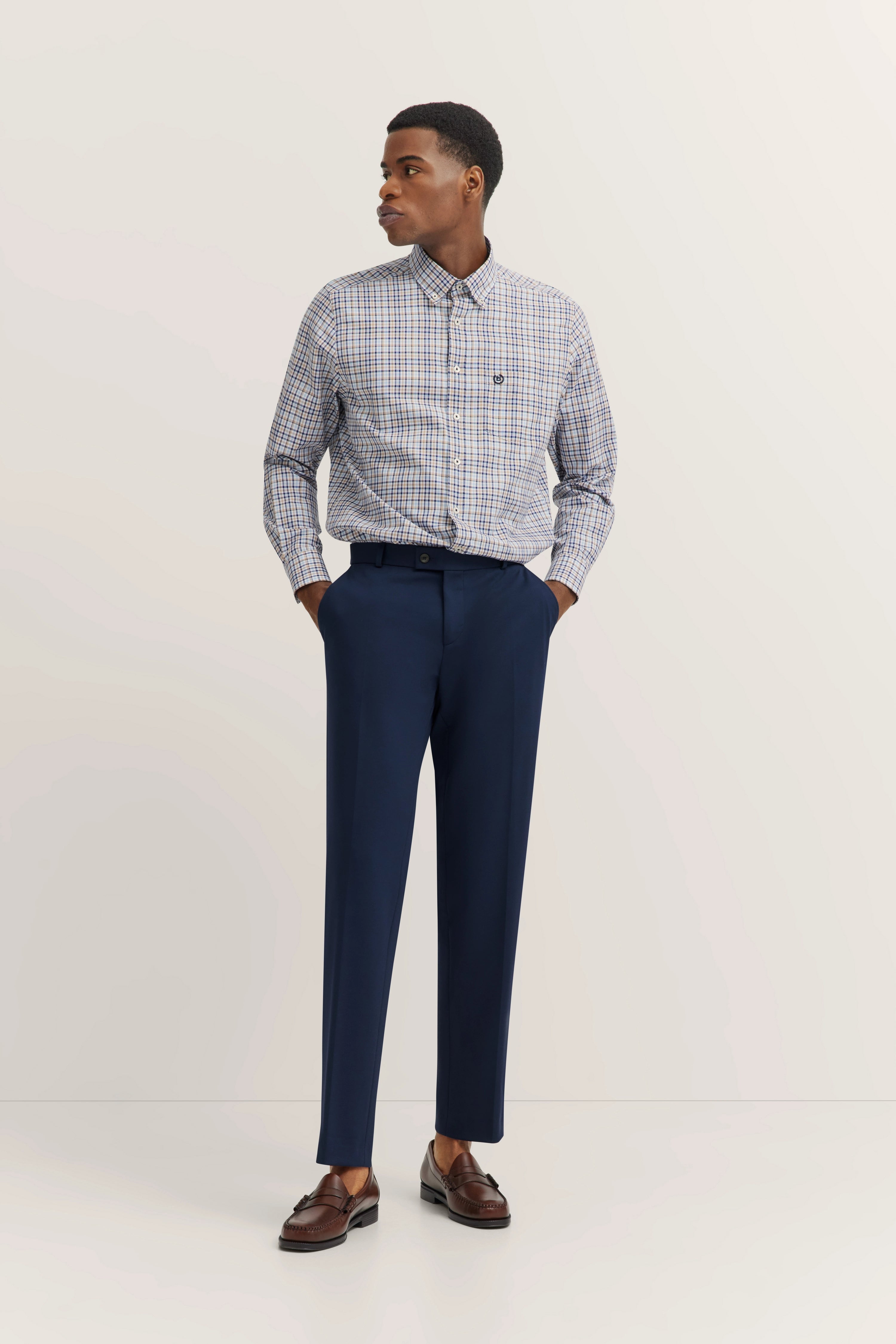 Model wearing a checked shirt and blue pants, hands in pockets, brown shoes by bugatti.
