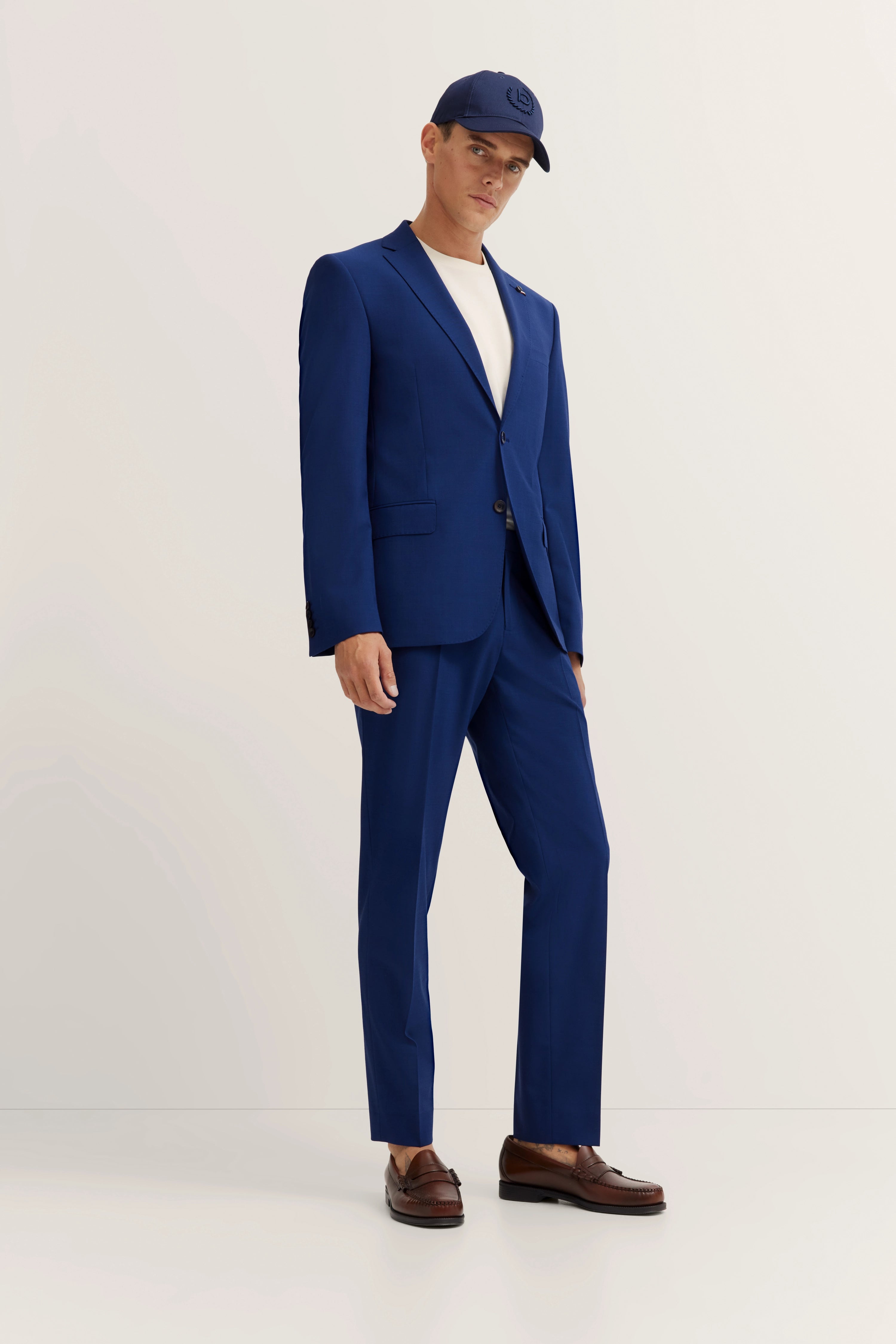 Model in blue bugatti suit, white shirt, brown shoes. Relaxed pose with blue cap.
