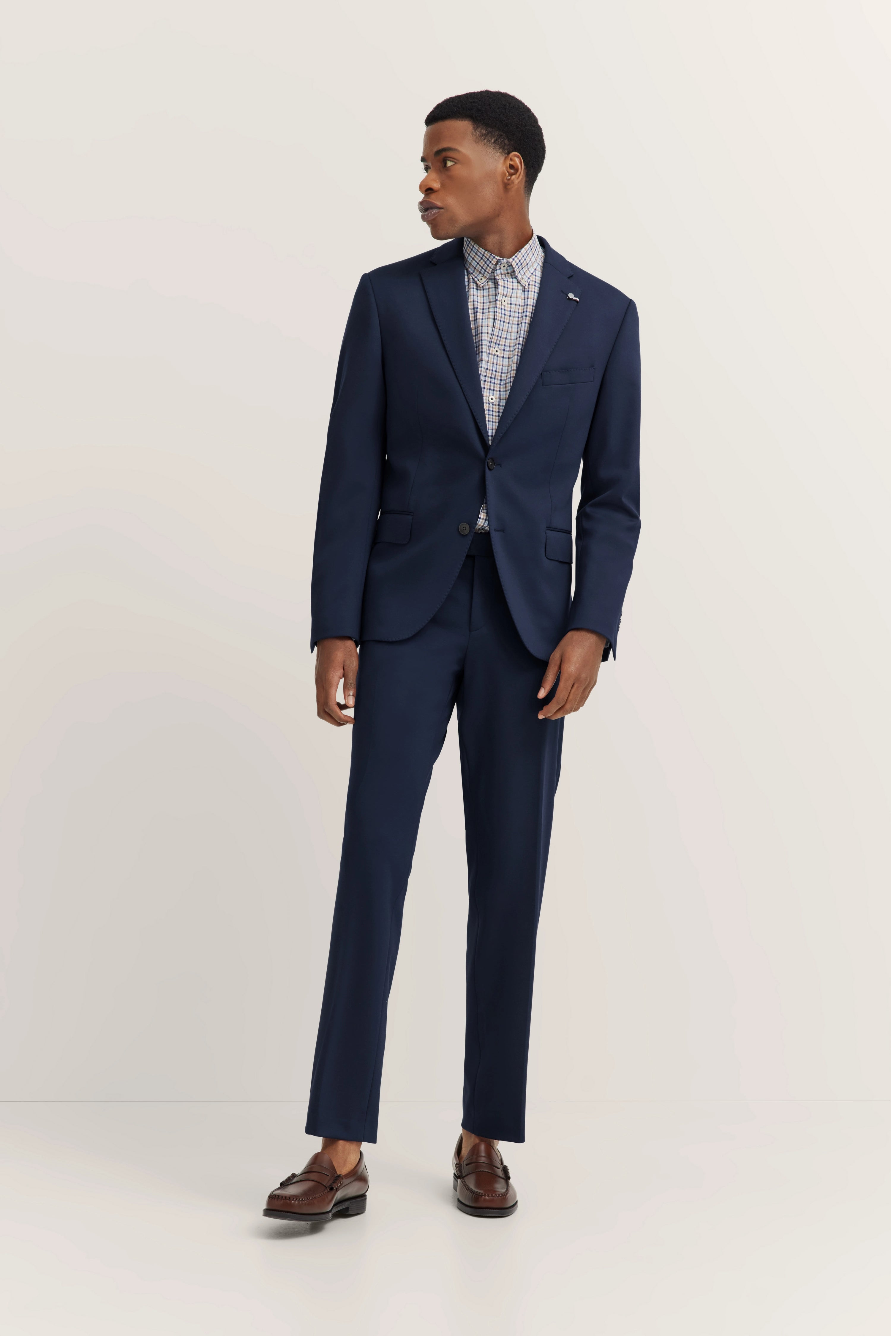 Model in dark blue suit, checkered shirt, brown shoes, posing sideways. Bust shot.