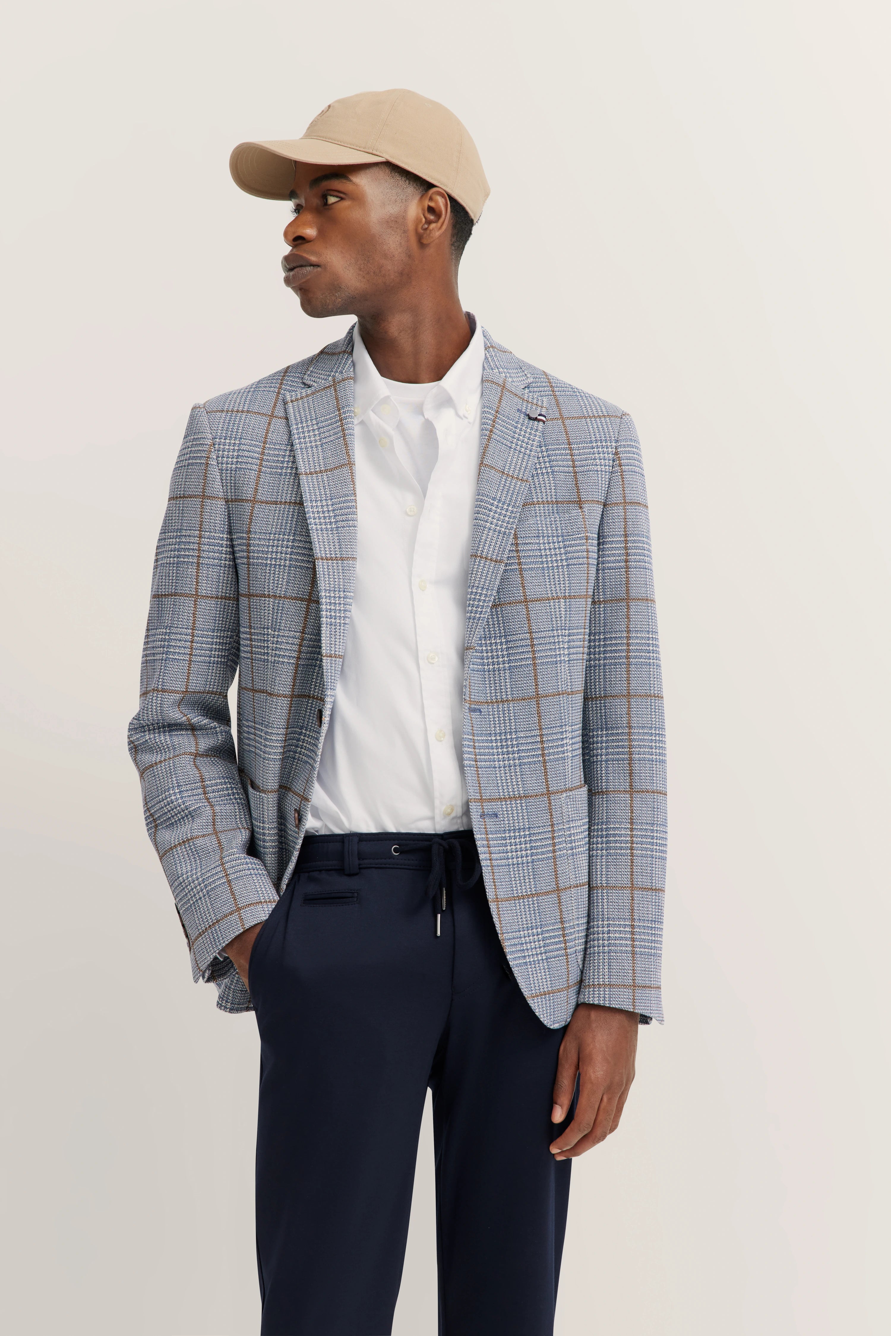 Model in beige cap, checkered bugatti blazer, white shirt and navy trousers, looking sideways.