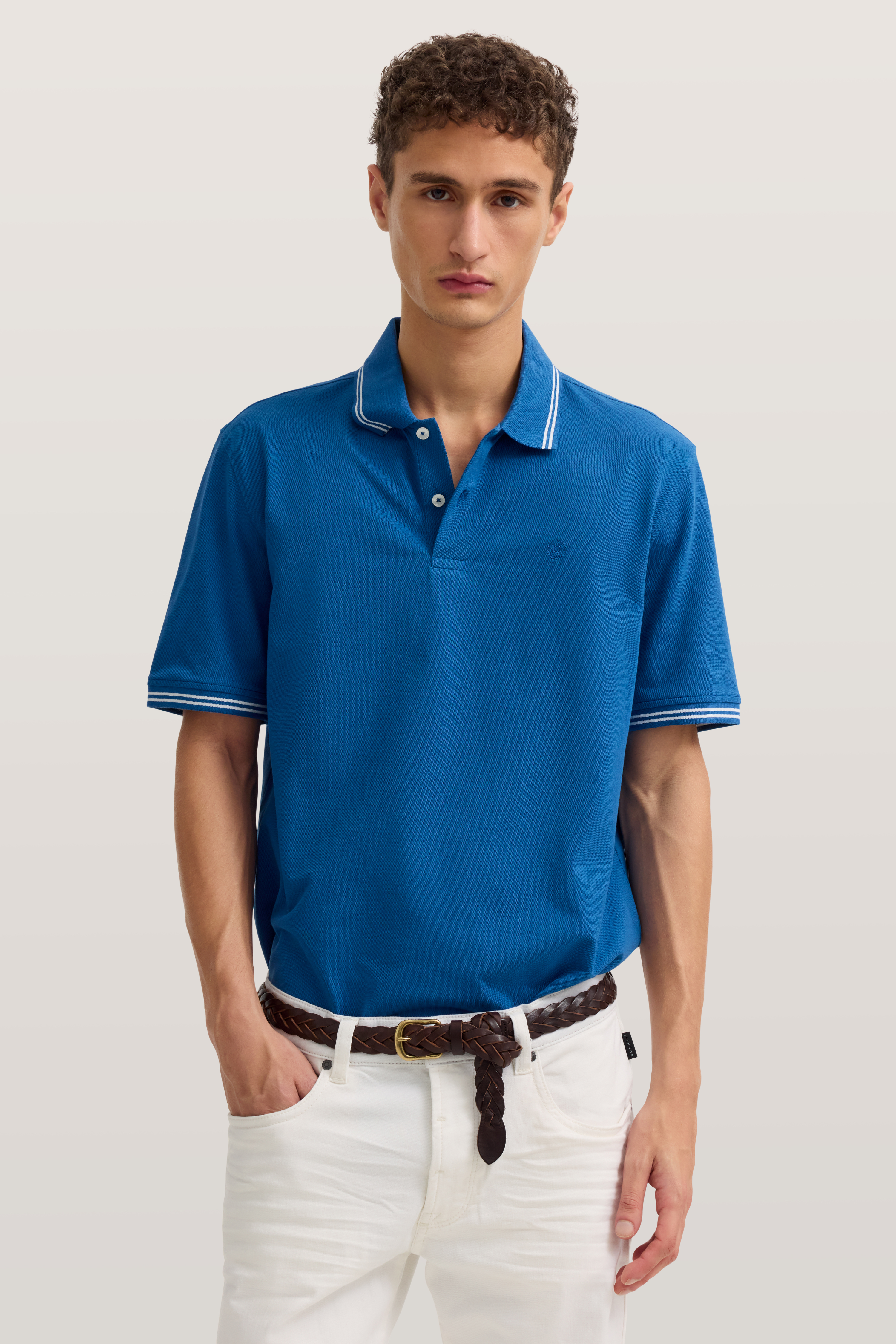 Model trägt blaues bugatti Polo und weiße Hose, lässige Pose, betont Stil und Gelassenheit.