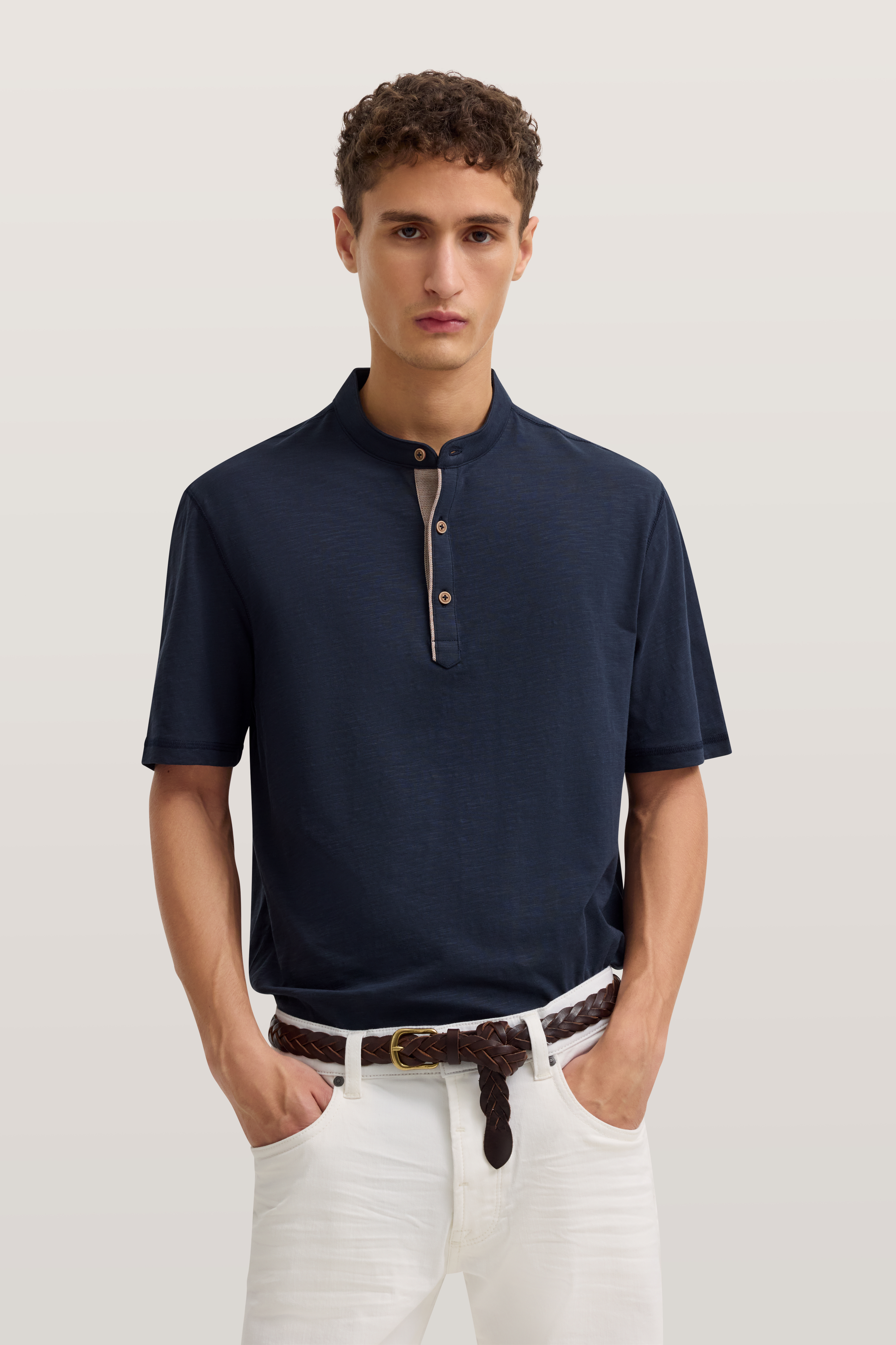 bugatti Modelbild: Dunkelblaues Shirt, Stehkragen, weiße Hose, Modell mit Händen in den Taschen, entspanntes Posing.