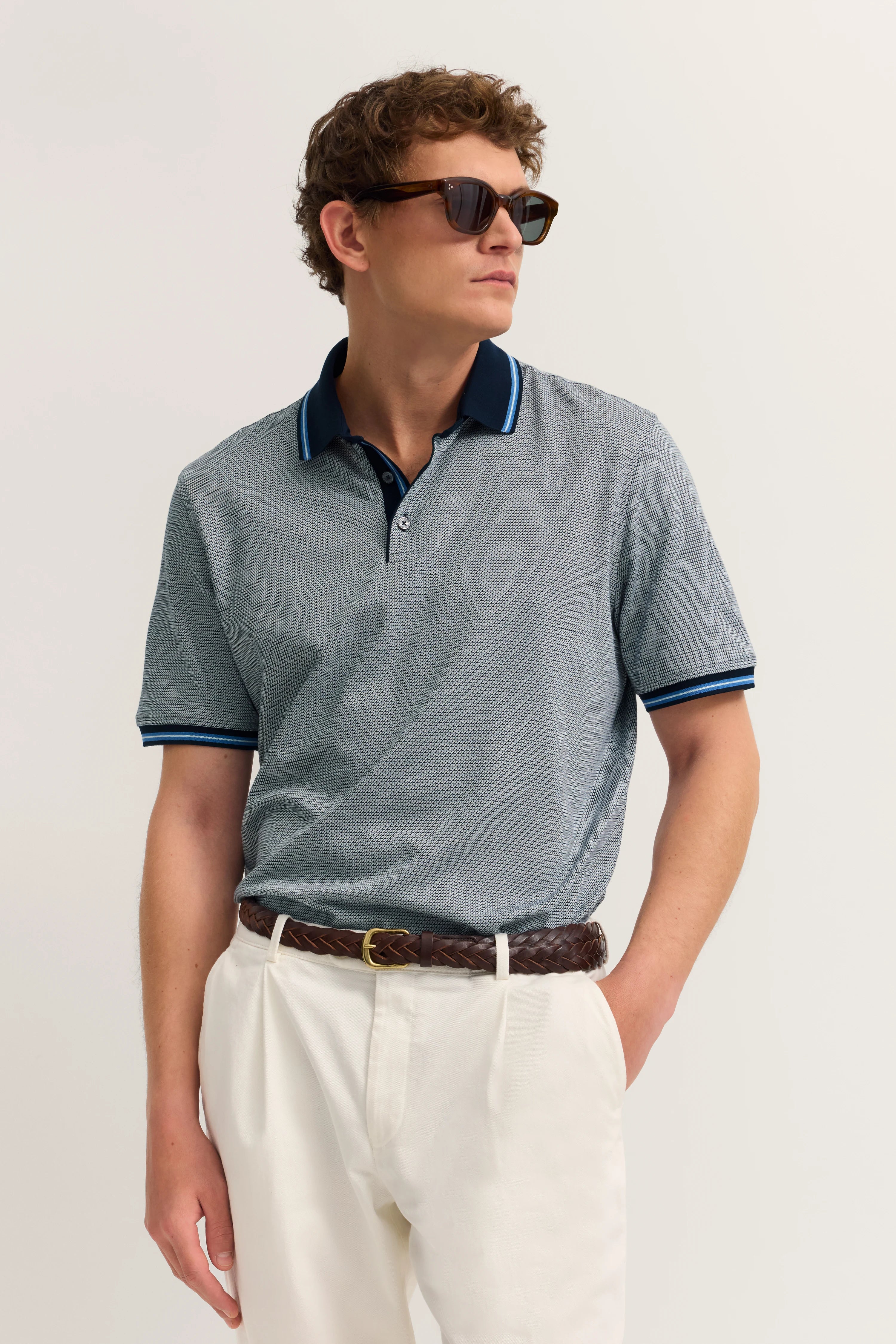 Bugatti Model in grauem Poloshirt, weißer Hose, braunem Gürtel, lässiger Blick nach rechts.